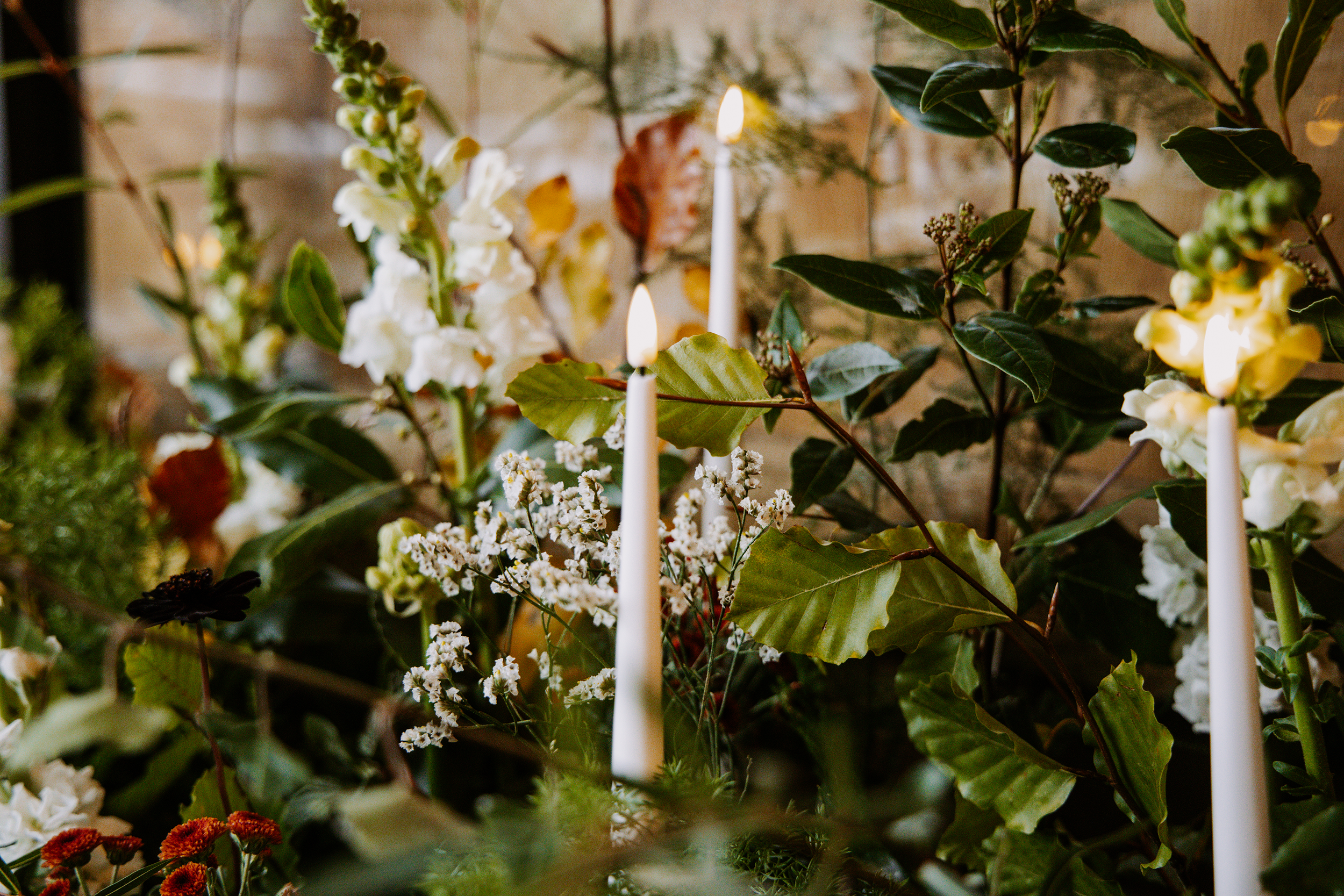 A close-up of a wedding floral arrangement with white candles, green leaves, white flowering plants, and small orange and dark flowers, set against a brick wall.