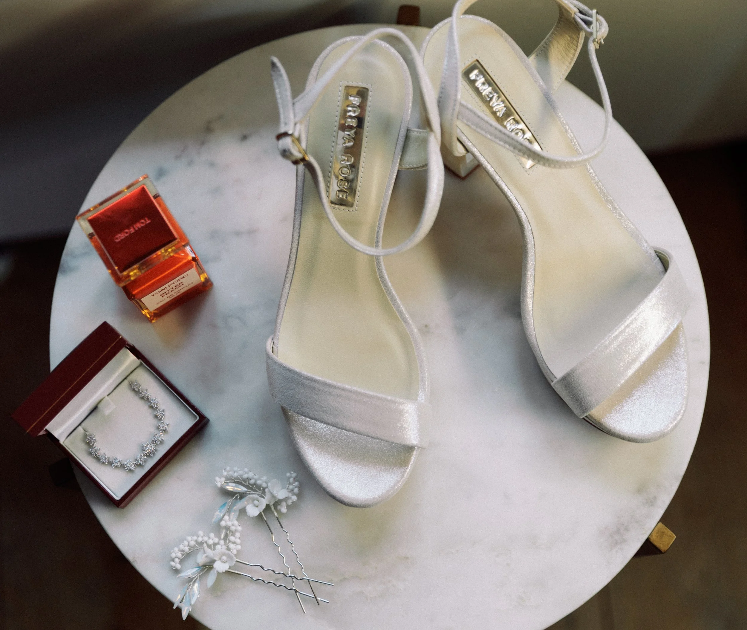 A white pair of high-heeled sandals with ankle straps, a red jewelry box with a diamond necklace, a pair of pearl hairpins, and a small red perfume box arranged on a round marble table.