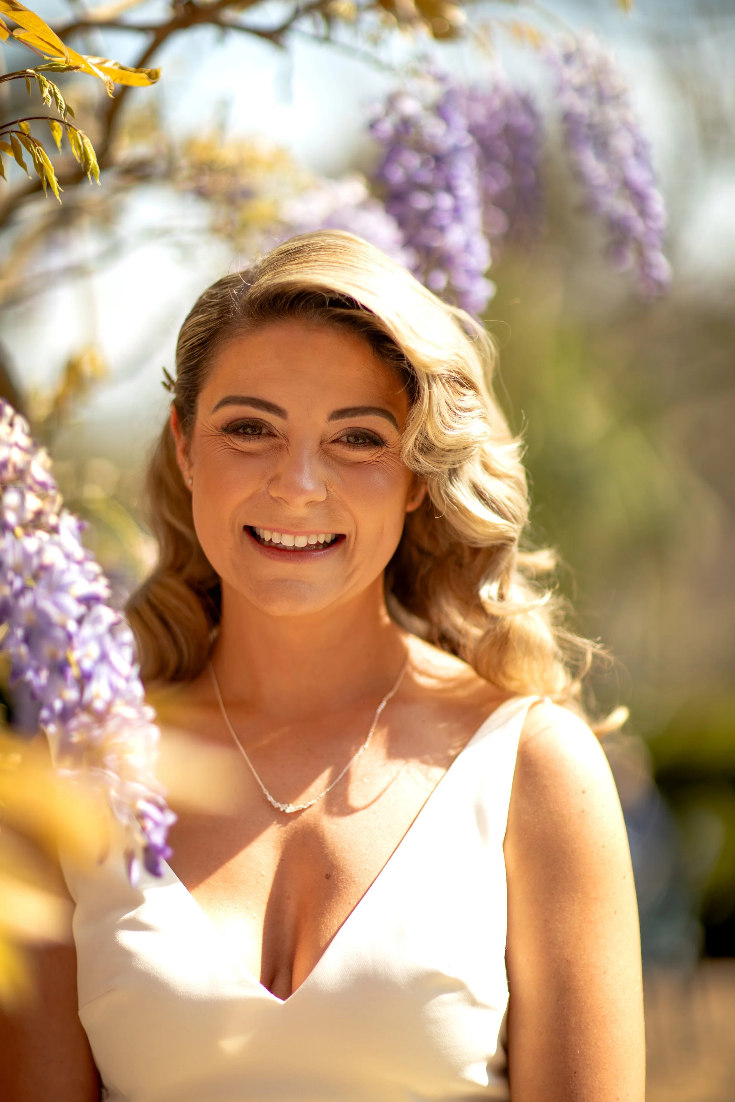 A woman in a white dress smiling outdoors surrounded by blooming purple flowers.