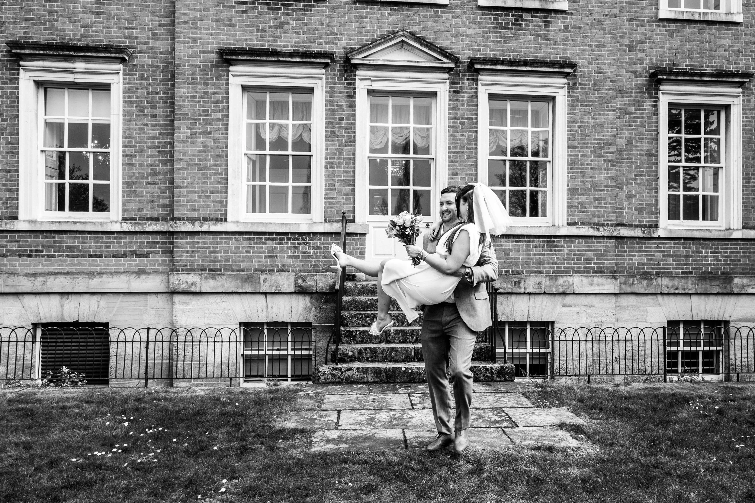 A couple dancing outside a brick building, with the man carrying the woman in his arms. The woman is holding a bouquet of flowers and wearing a wedding dress and veil, while the man is in a suit. The scene is cheerful and romantic.