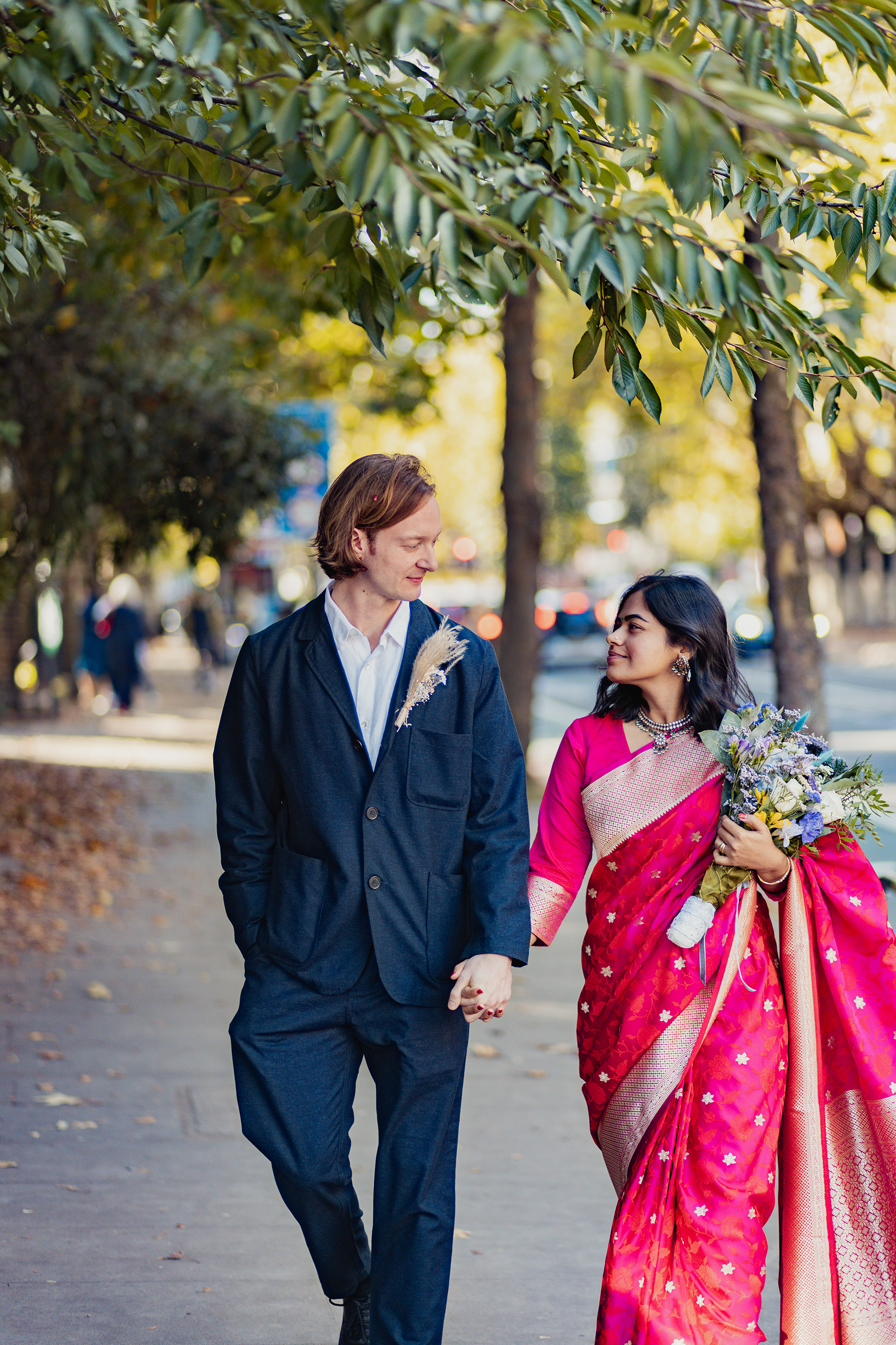 Couple walking hand in hand outdoors, with the woman holding a bouquet of flowers. The woman is dressed in a pink traditional outfit, and the man is in a dark blue suit, both smiling at each other.