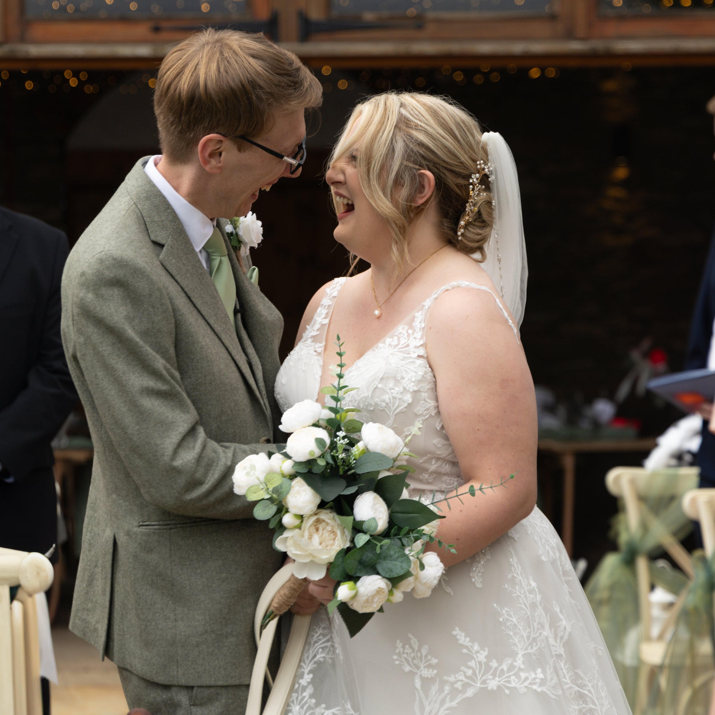 A bride and groom laughing and holding hands at their wedding ceremony, with the bride holding a bouquet of white flowers and greenery.