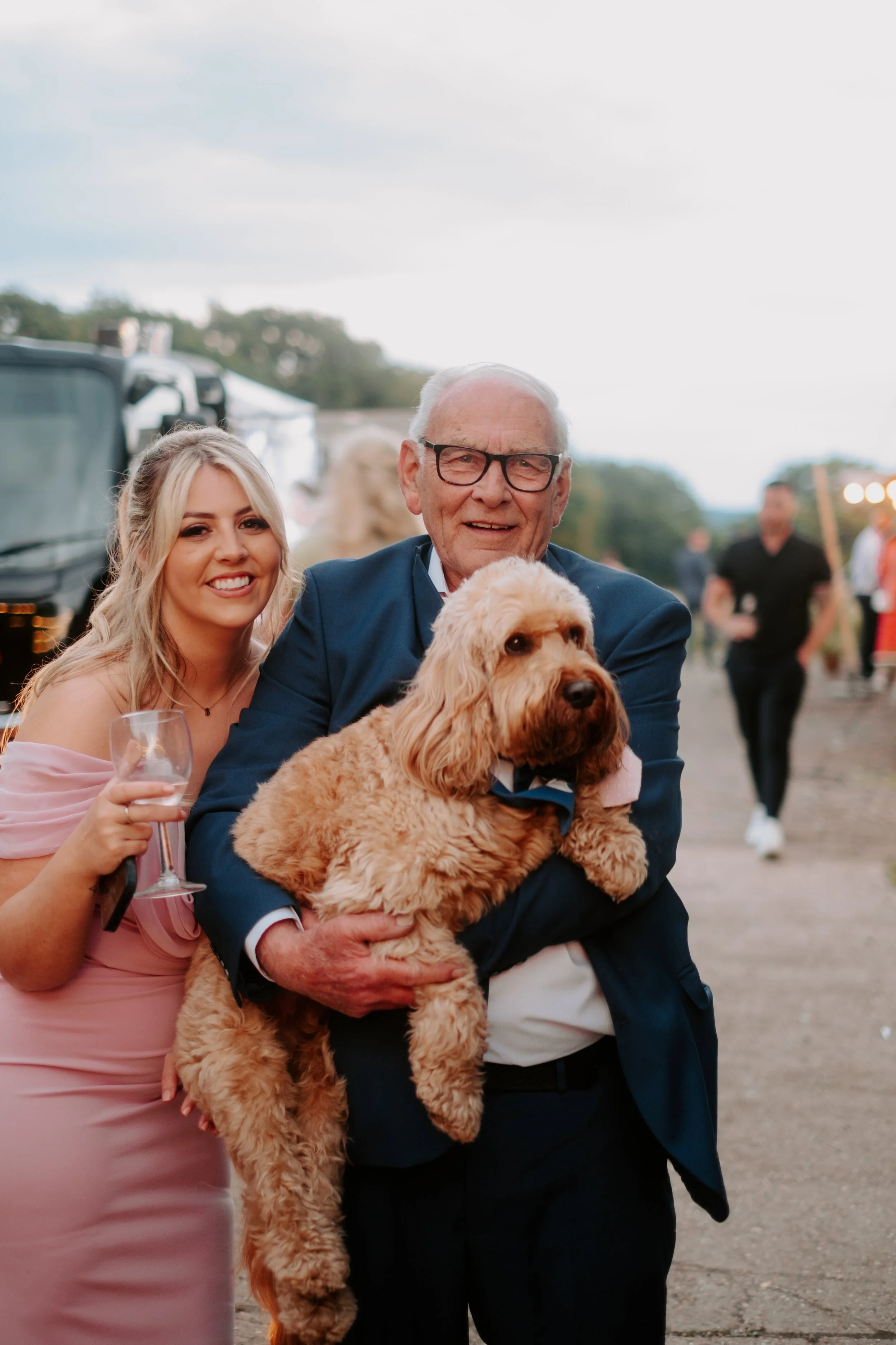 A young woman in a pink dress holds a glass of wine and smiles next to an elderly man in a navy suit holding a large curly-haired dog during an outdoor event at dusk.