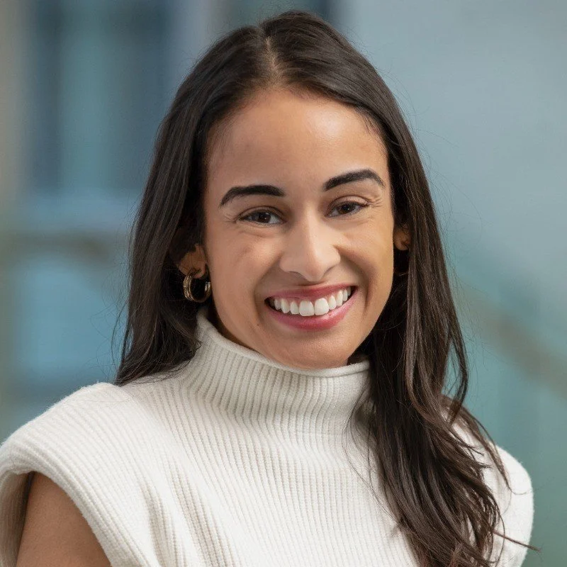 Young woman with long brown hair smiling, wearing a white turtleneck sweater and hoop earrings, indoors with blurred background