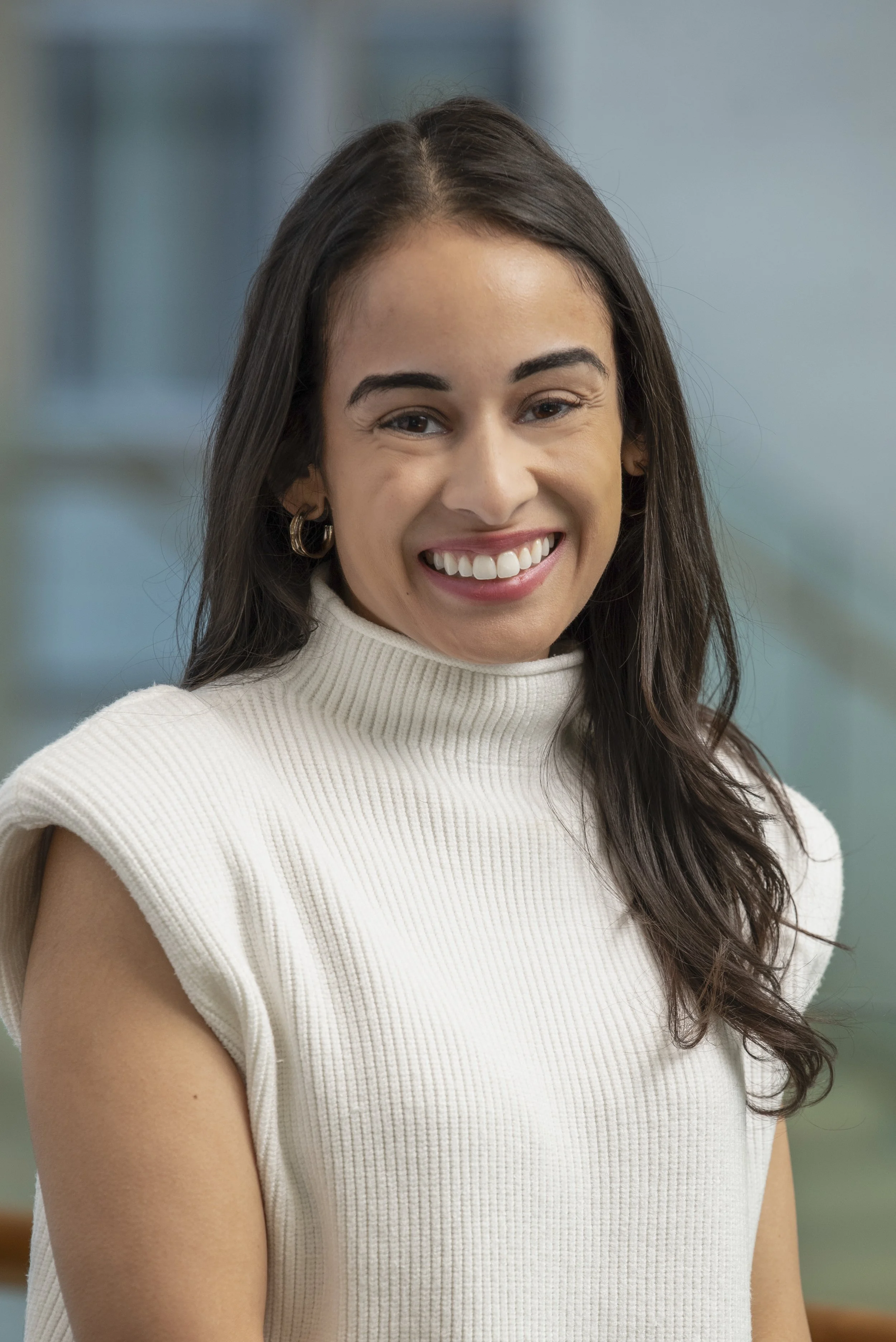 A woman smiling and wearing a cream sleeveless turtleneck, with long dark hair and hoop earrings, indoors with a blurred background.