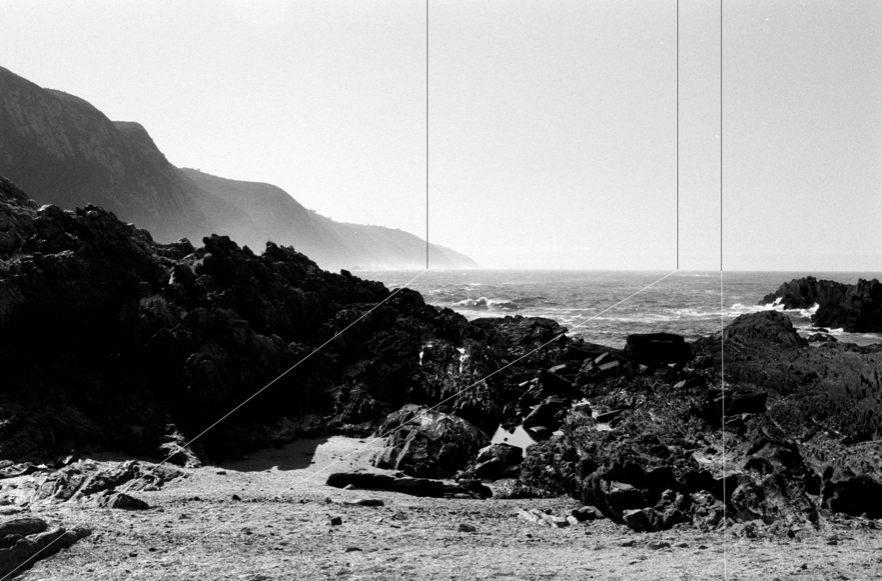Black and white photo of a rocky beach with cliffs in the background and ocean waves near the rocks.