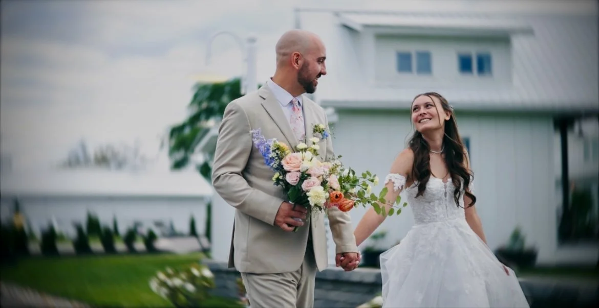 A groom holding a bouquet of colorful flowers and a bride in a white wedding dress holding hands and smiling at each other outside in front of a house.Bride and groom holding hands in a cinematic wedding film by SBS Wedding Films