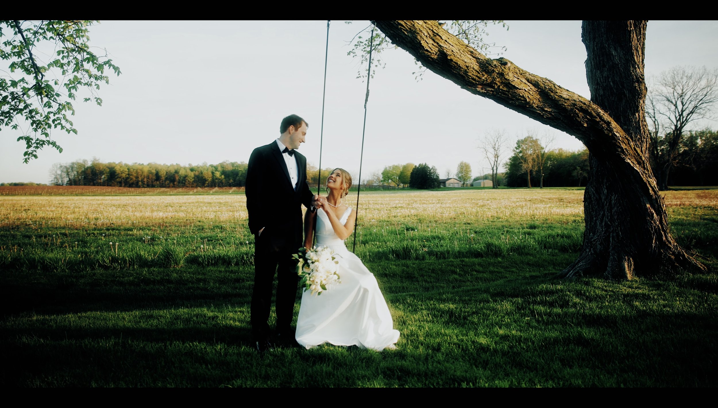 A bride and groom standing under a tree on a grassy field, wedding scene, couple smiling at each other, bride holding a bouquet of white flowers, groom in a black tuxedo, sunny day.