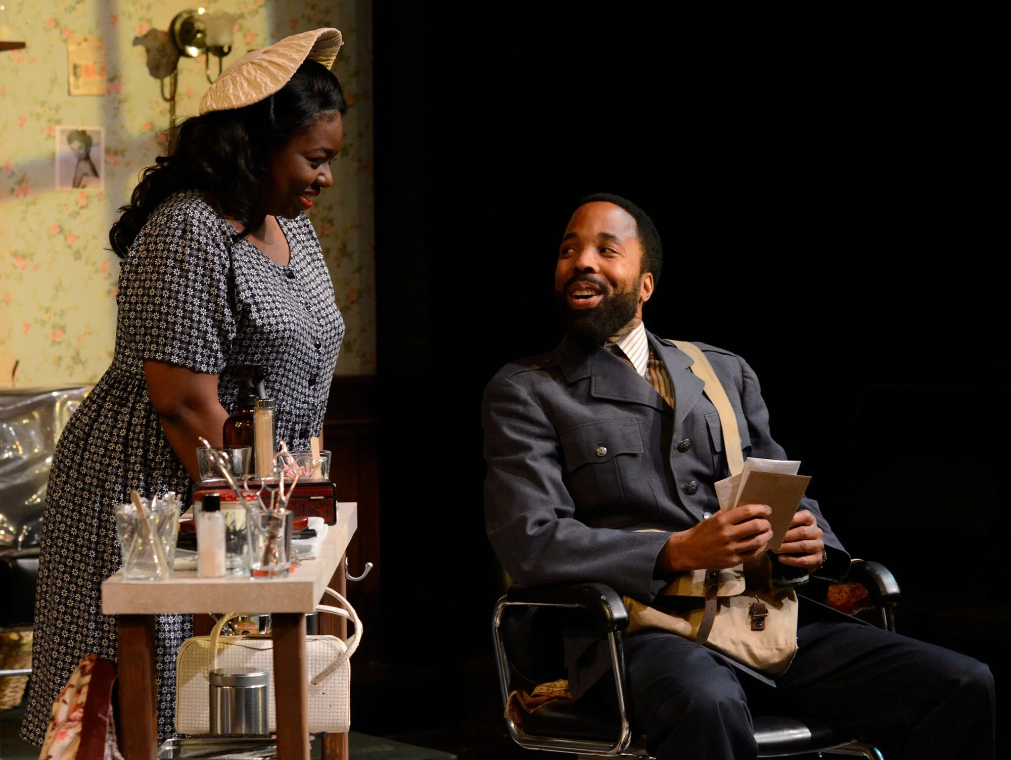 A woman and a man in costume perform on stage. The woman stands behind a makeup table, leaning toward the man who sits in a chair holding a paper. The set appears to depict a vintage or historical setting.