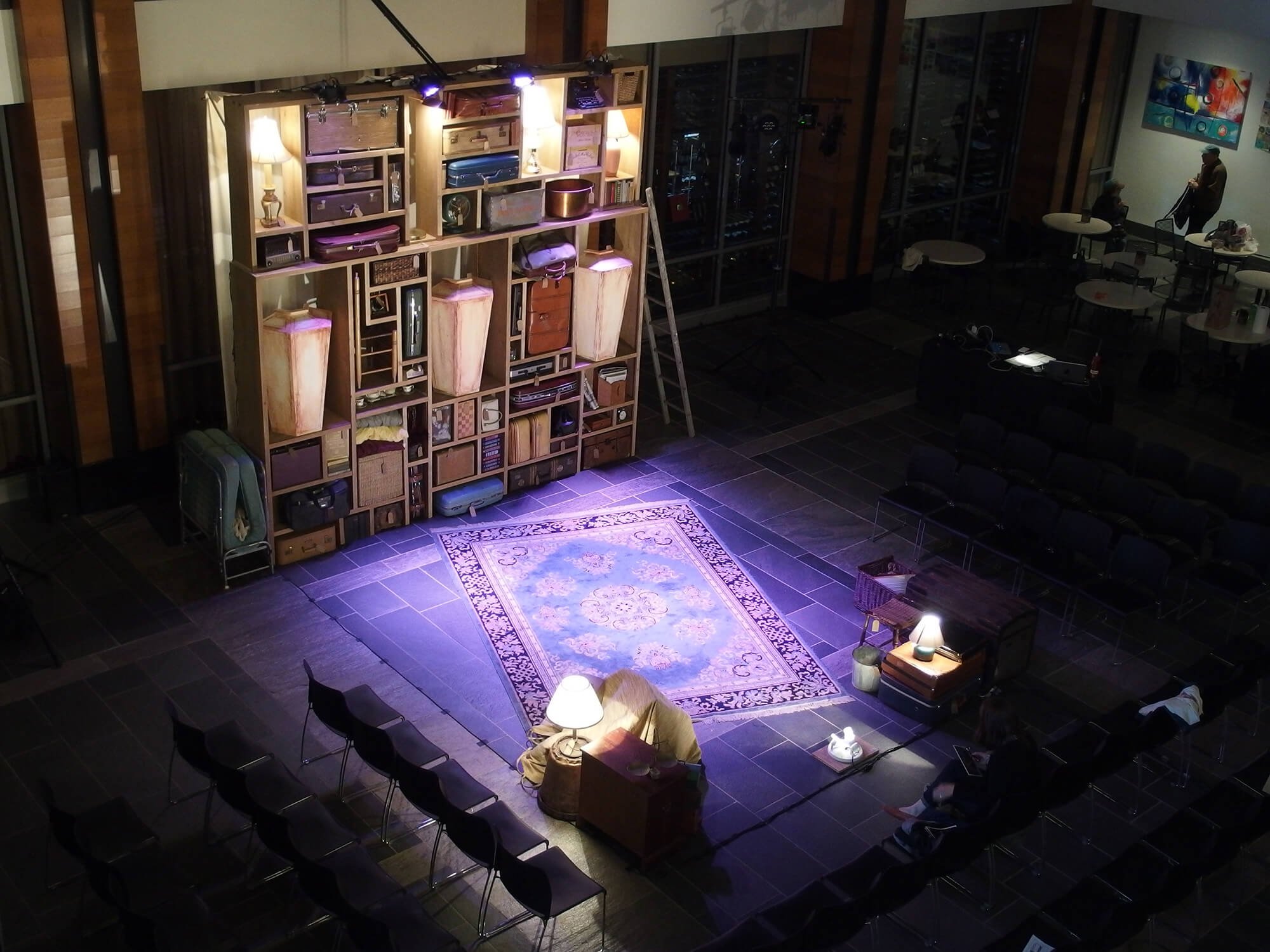 Empty theater or event space with a small stage area illuminated by lamps and a spotlight, surrounded by chairs arranged in a semi-circle. A large wooden shelving unit, partially lit, holds luggage, boxes, and decor, with a ladder leaning against it.