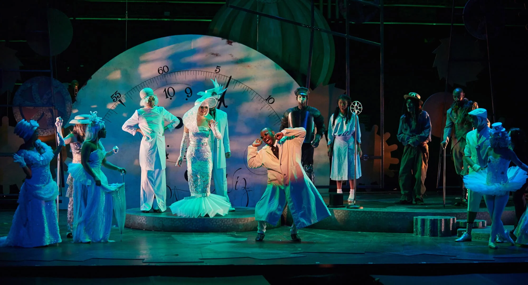 Theater stage with actors in colorful costumes, some in wedding attire and others dressed as characters from a story, with a large clock background.
