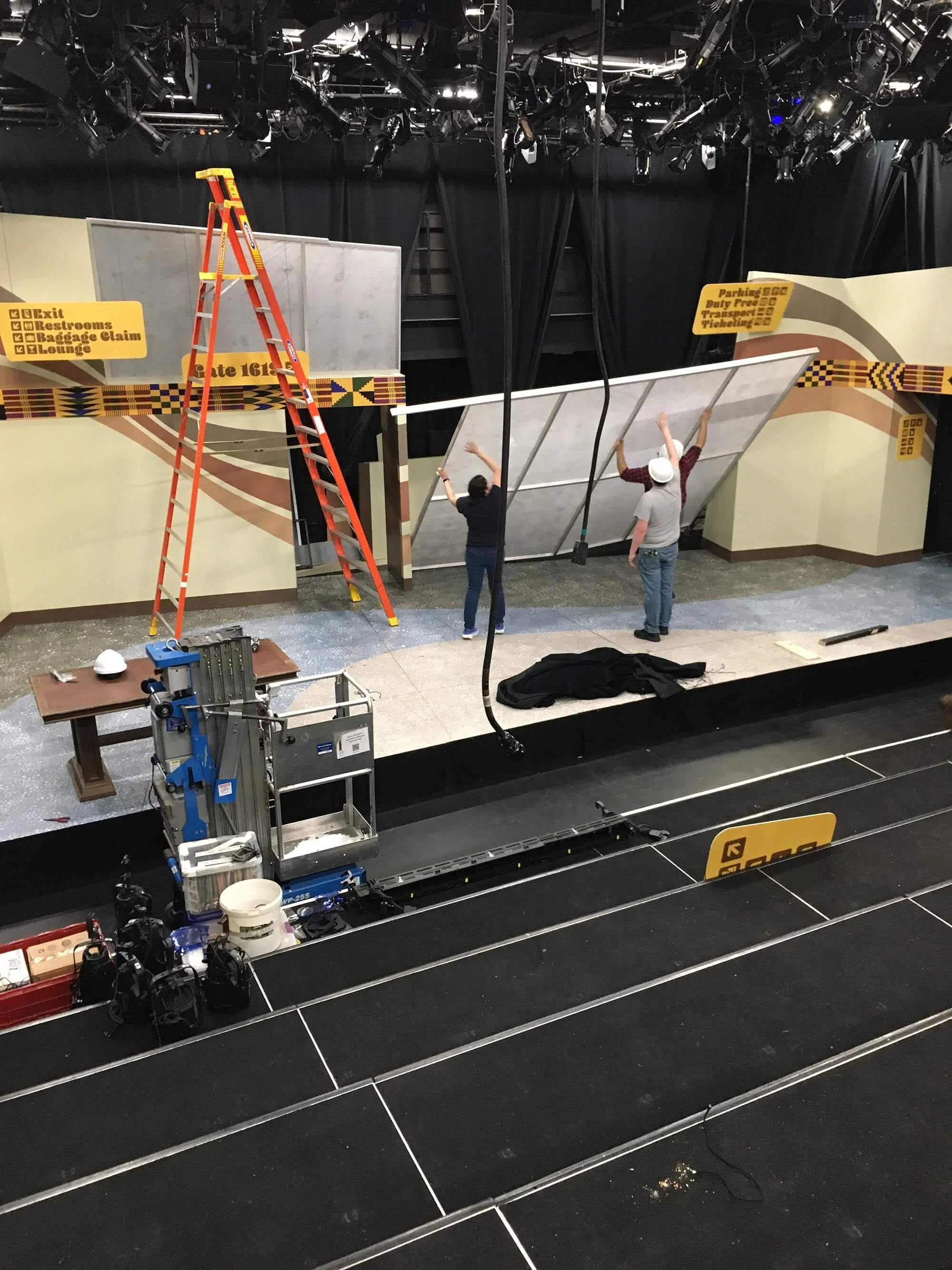 Workers setting up a stage with a painted backdrop, ladder, and equipment, with signs indicating directions in a behind-the-scenes area.