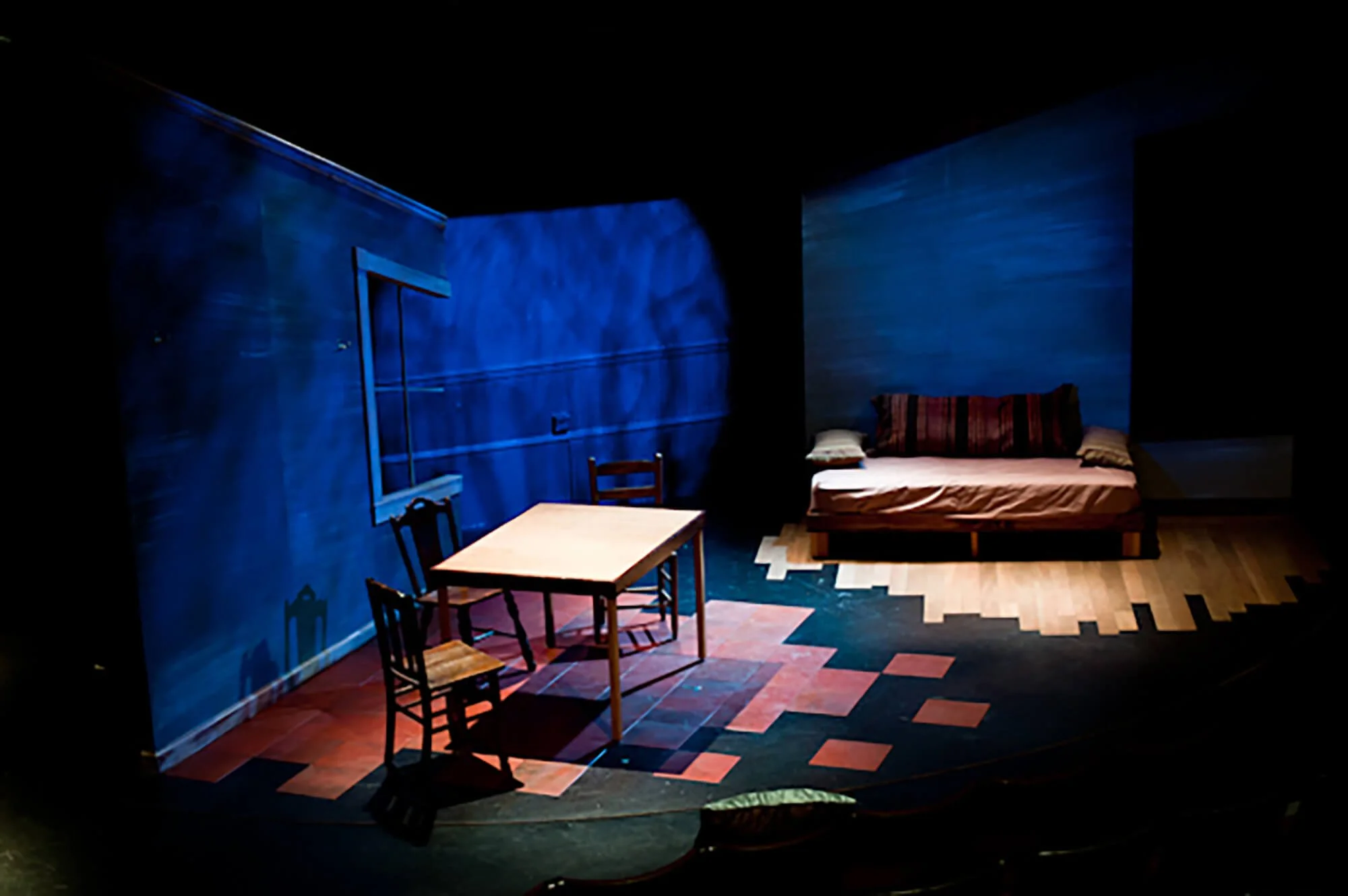 Theater stage set depicting a blue wall, a small wooden table with three chairs, and a bed with pillows on a wooden floor, illuminated with theatrical lighting.