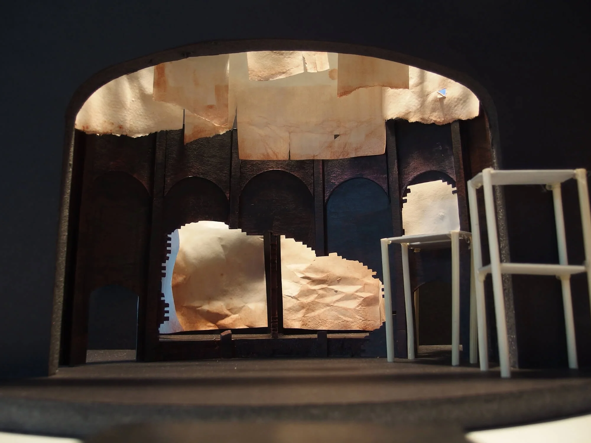 A miniature architectural model with black and white structural elements, including archways and stairs, inside a darkened space with tissue paper decorations.