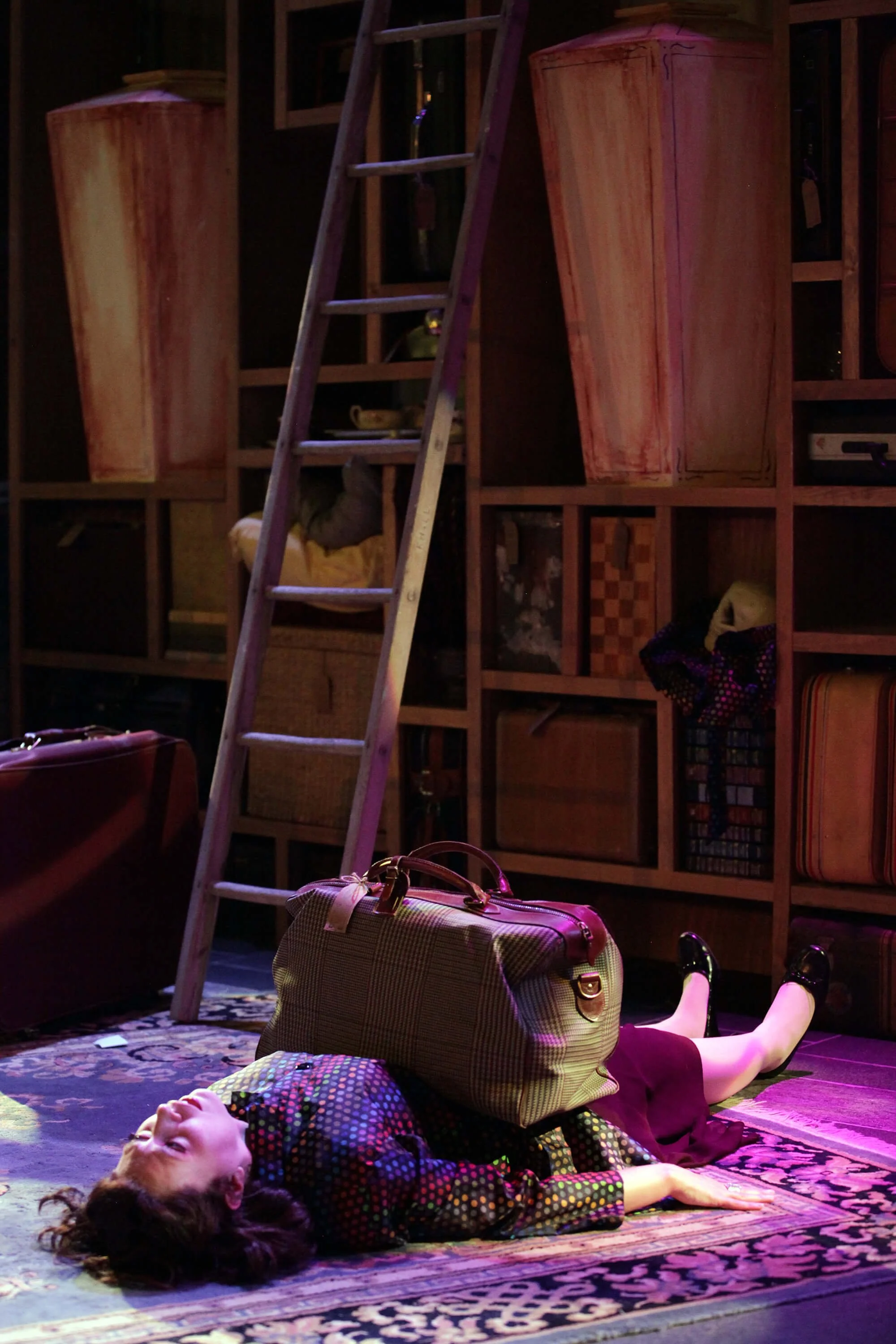 Woman lying on the floor with a plaid suitcase on her stomach, in a room with a ladder and shelves filled with bags and items.