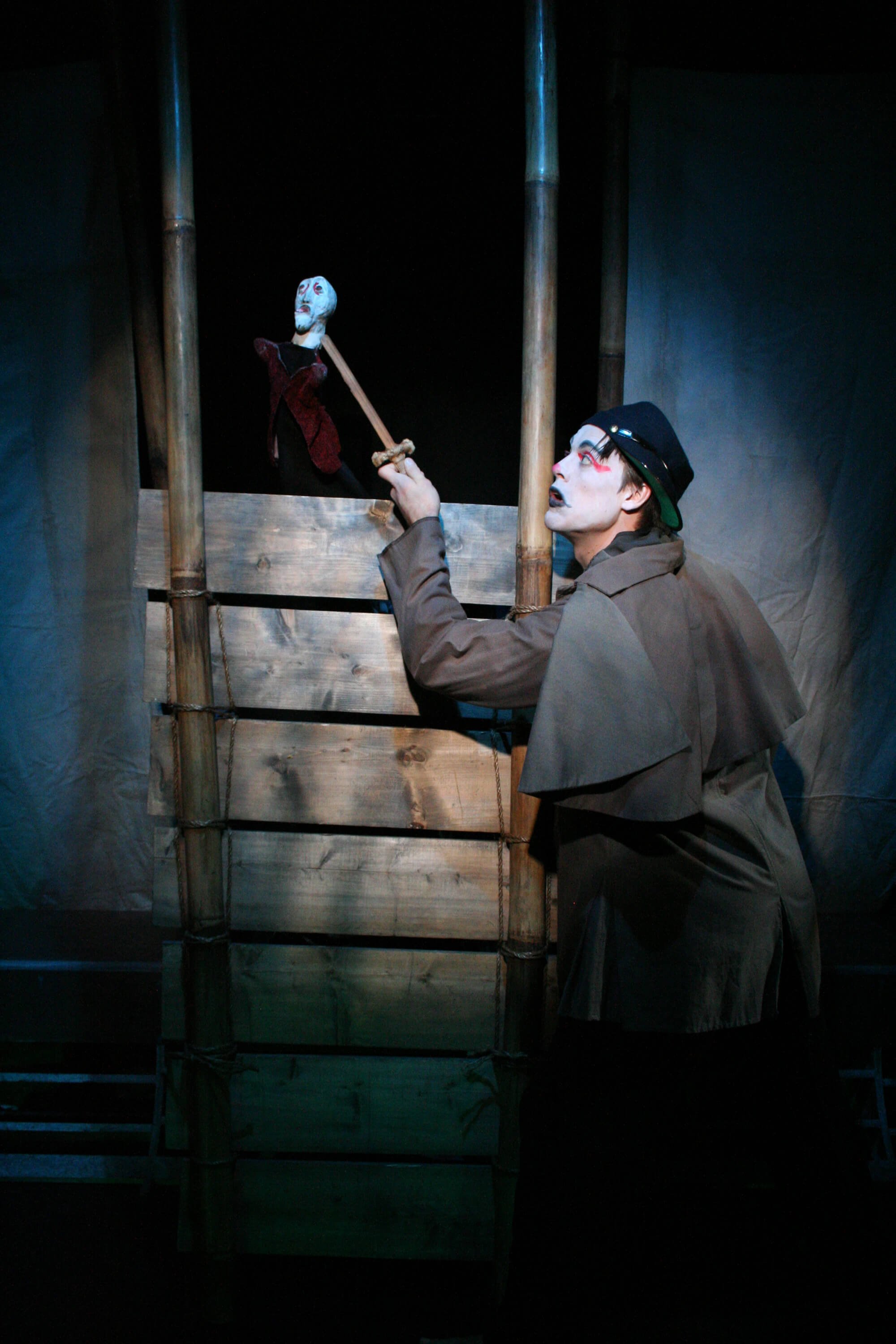 A mime performer dressed in brown costume and painted face, holding a stick and looking up at a puppet on a small wooden stage, with a dark background.