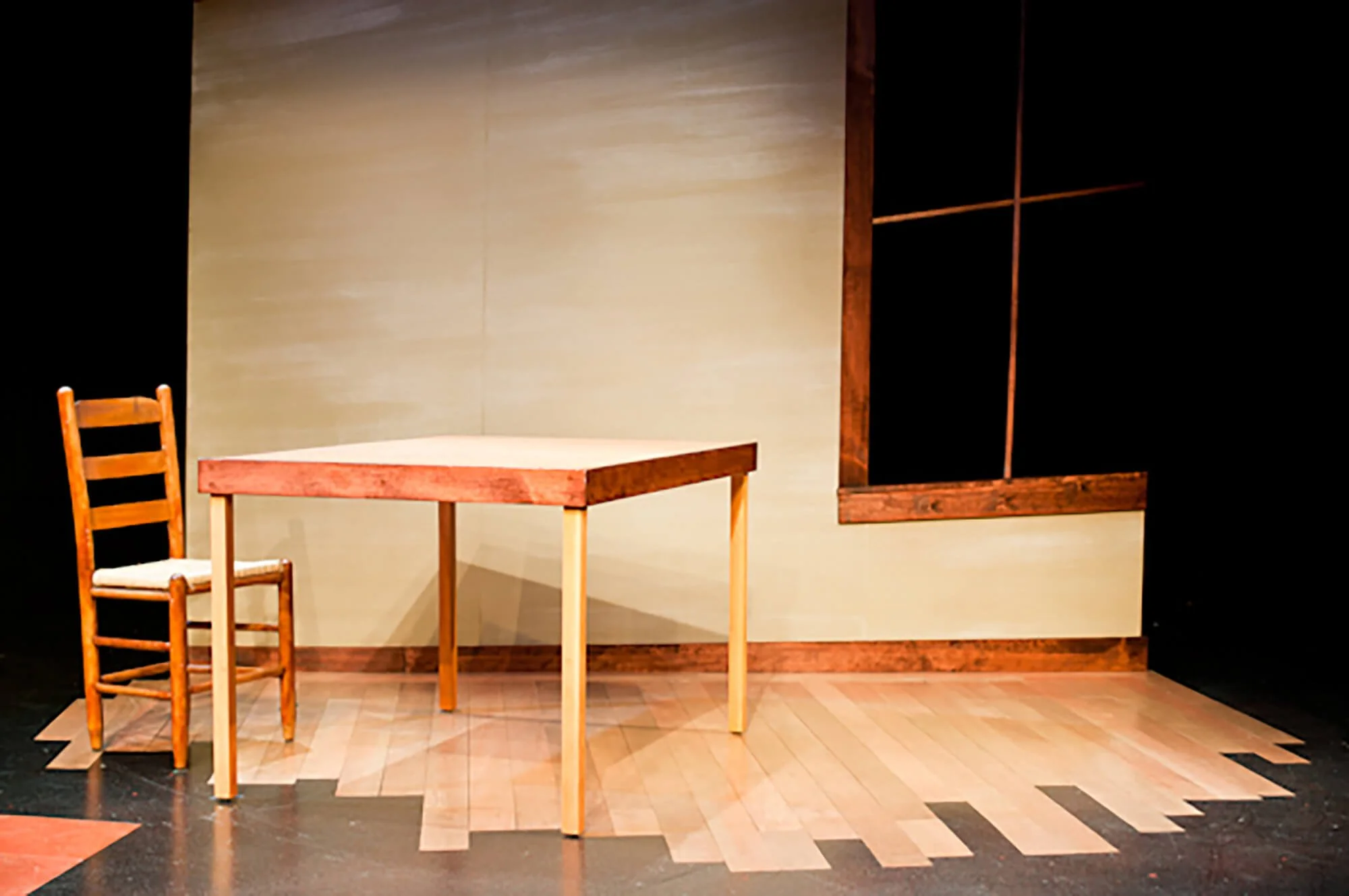 Stage set with a wooden chair, a wooden table, and a beige wall background with a window frame.