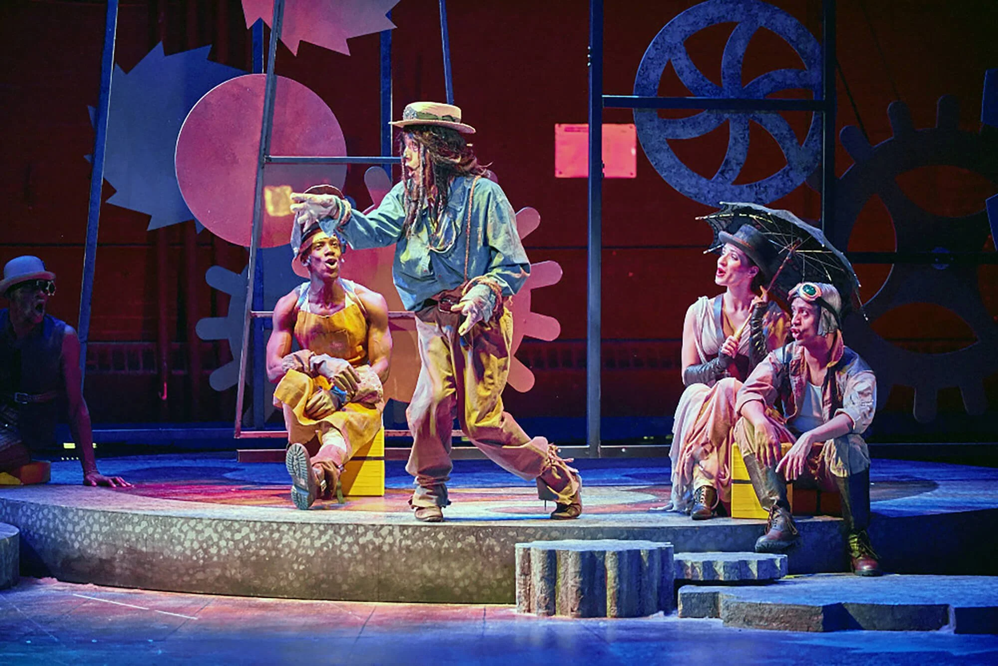 Theater scene with performers on stage surrounded by colorful gear and machinery props, with one performer standing and pointing while others sit and listen.