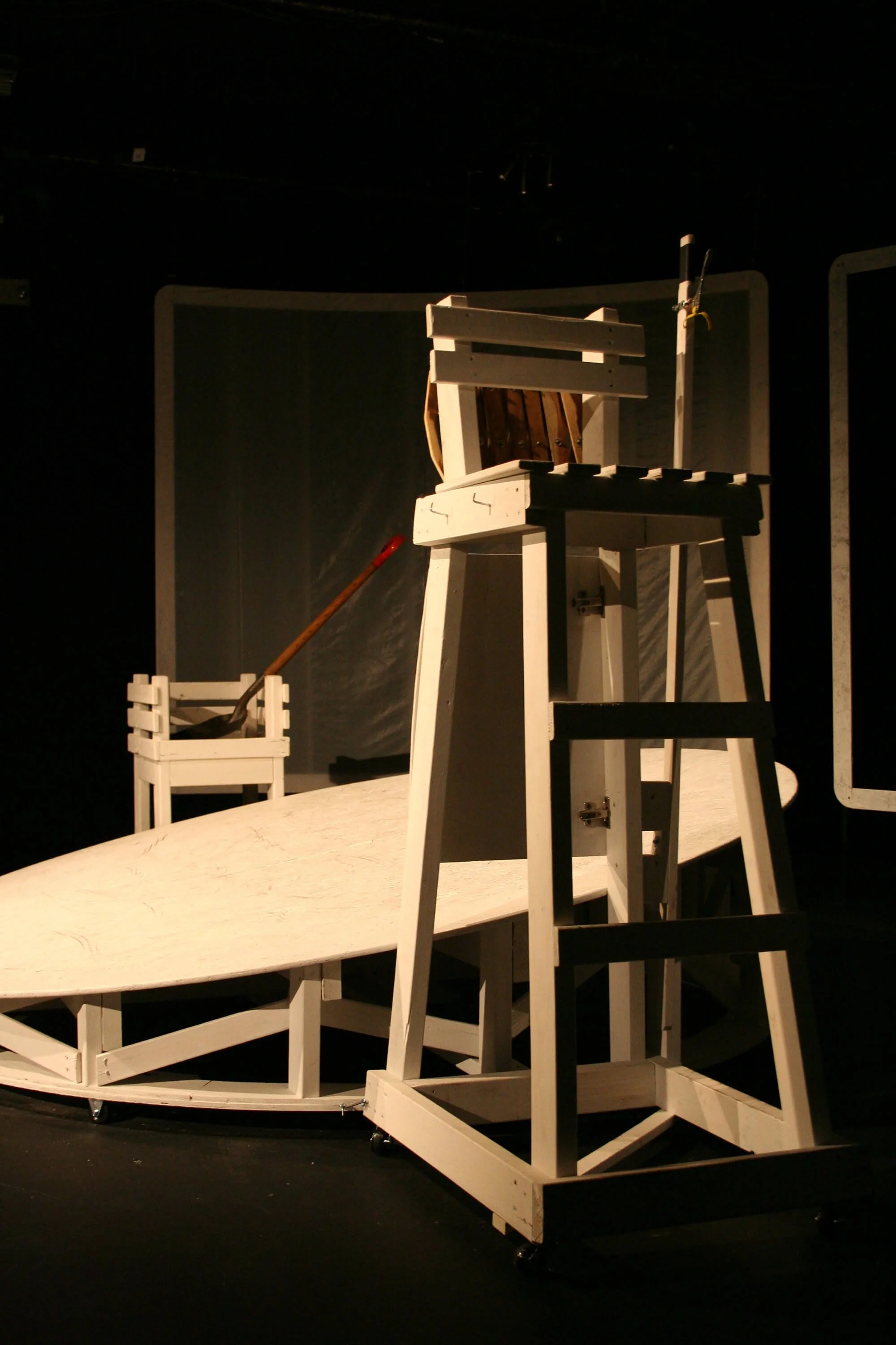 Stage prop resembling a small boat with a wooden deck, a chair, a small platform, and a potted plant, all made of white painted wood, set in a dark theater or studio setting.