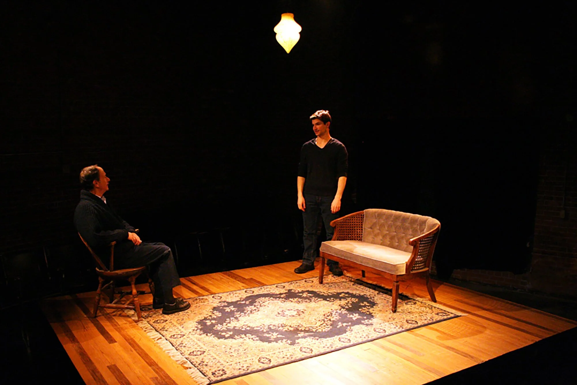 A theater scene with two men, one seated on a wooden chair and the other standing, in a dimly lit room with a vintage rug, a sofa, and a hanging light fixture.