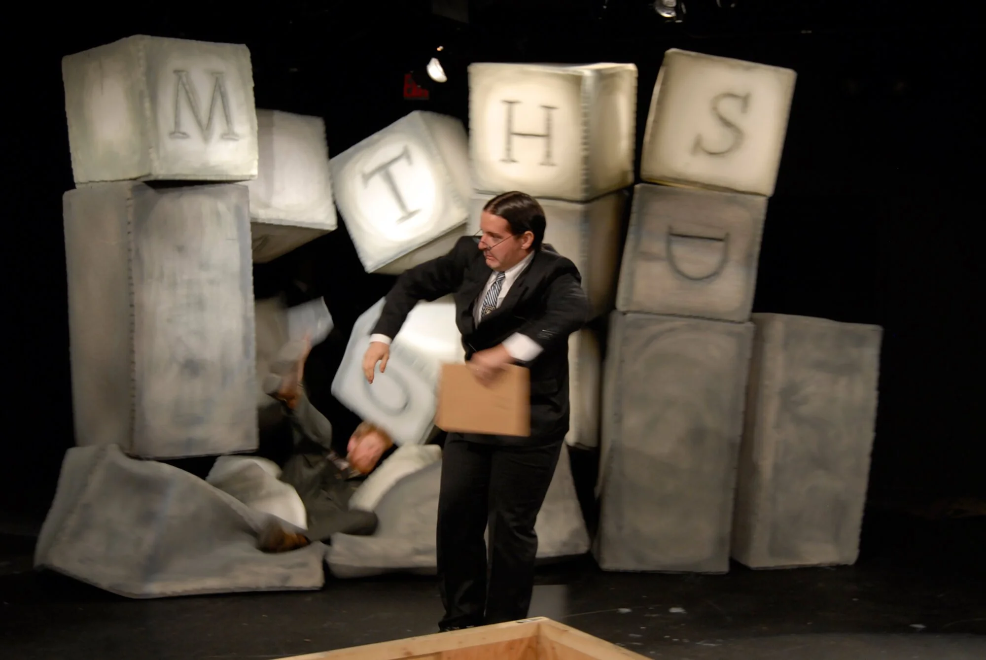 A man in a suit and tie appears to be collapsing or falling into a pile of large, soft blocks resembling baby blocks, with some blocks still stacked and others toppled. The background is dark, suggesting a stage or theatrical setting.