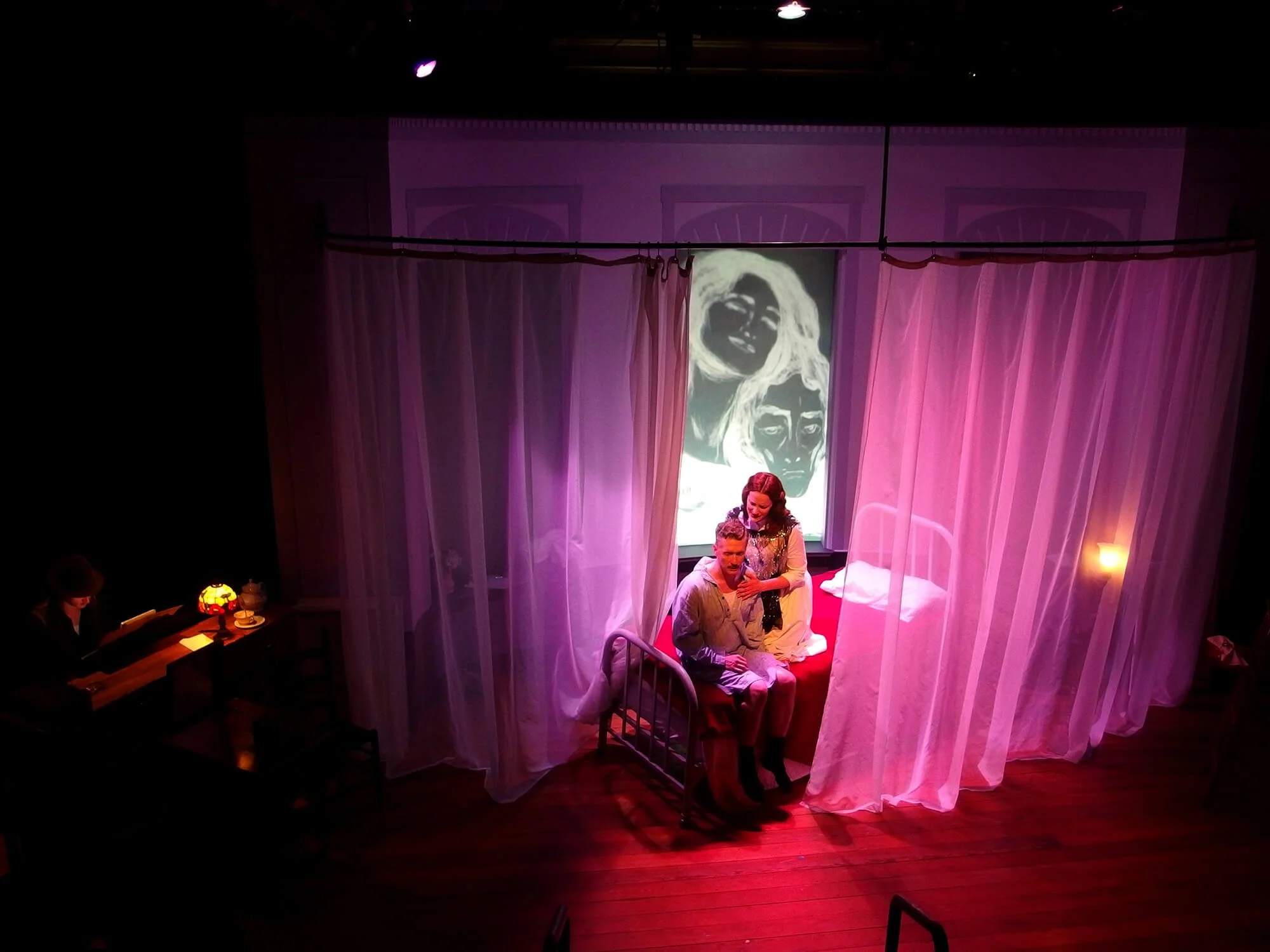 A stage scene with a bed draped in white curtains, a man and woman sitting on the bed, and a backdrop with a black and white portrait of two women. The woman is standing behind the man, holding his shoulders. The scene is lit with pink and purple lighting.