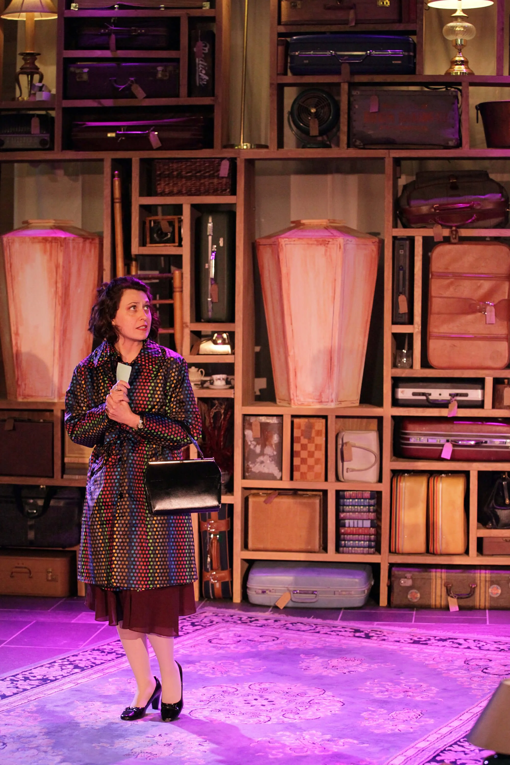 A woman with curly dark hair wearing a colorful polka dot coat and holding a black handbag, standing in front of a large wooden shelving unit filled with vintage suitcases and decorative items, illuminated with purple lighting.