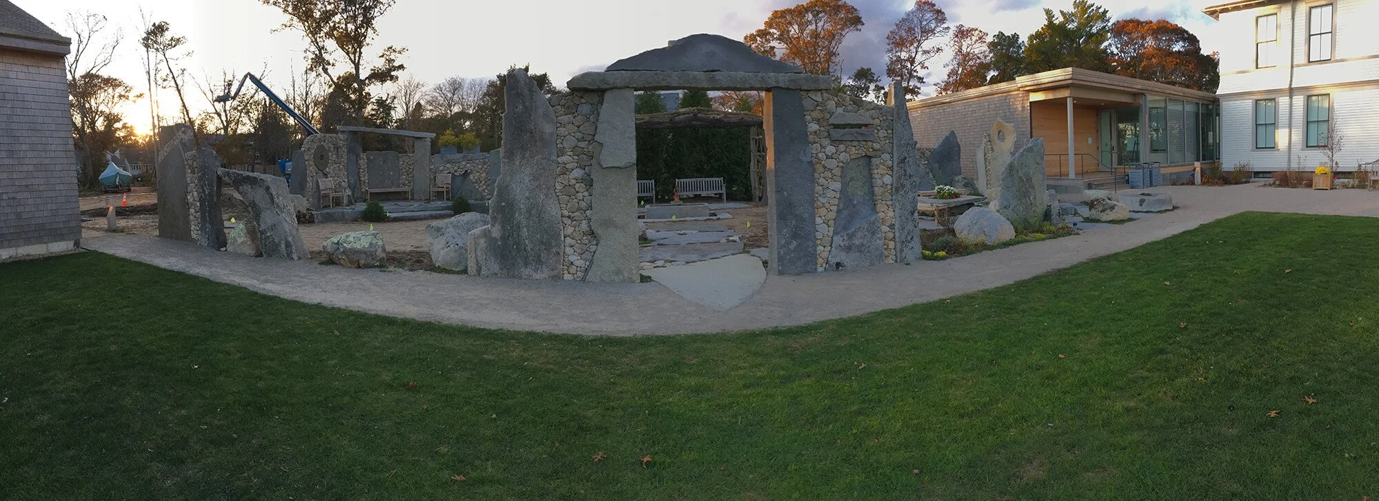 A stone and gravel outdoor structure with large rocks arranged vertically and a stone archway, surrounded by a concrete walkway, benches, and a grassy area, with a modern building with large windows and trees in the background during sunset.