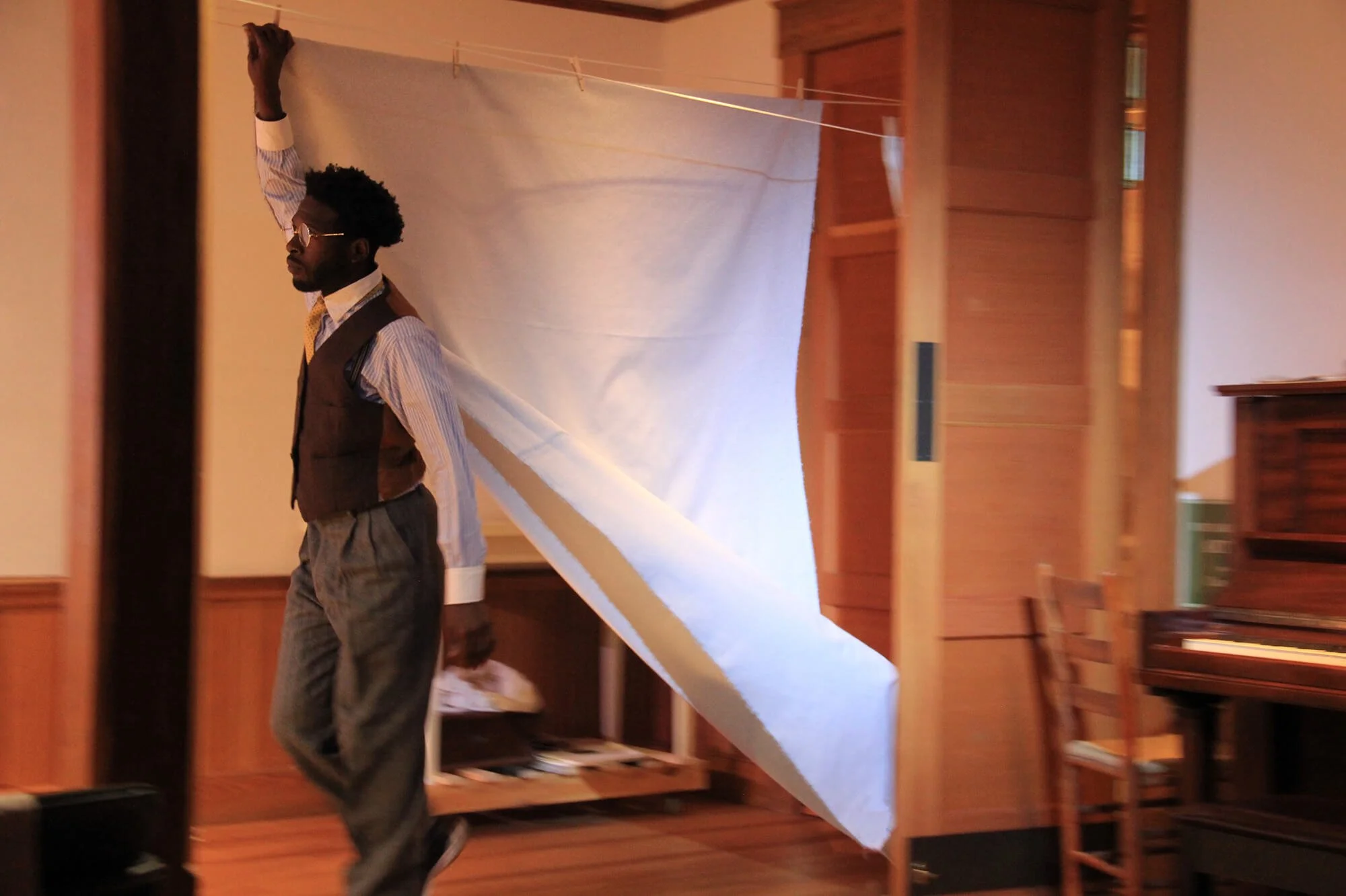 A man dressed in vintage style clothing with a vest, striped shirt, and sunglasses, walking past a makeshift photography backdrop made of a white sheet hanging from a clothesline in a wooden-paneled room.