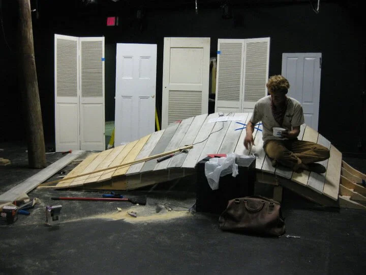 A person sitting cross-legged on a stage with wooden panels, surrounded by tools and supplies, with several white folding doors and a black wall in the background.