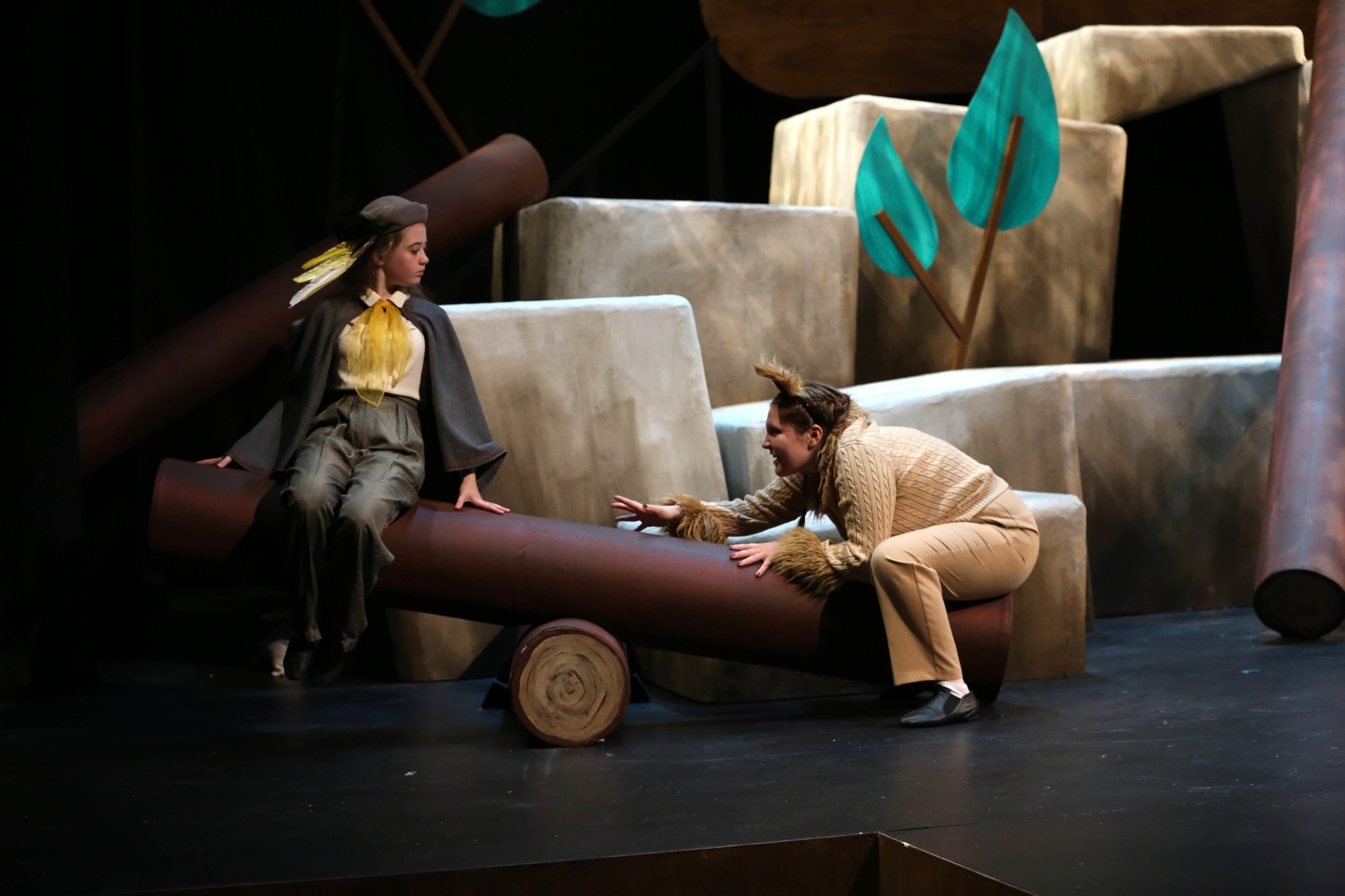Two young women dressed in costumes performing on a stage with a set resembling a forest, featuring large painted trees and log structures. One woman is sitting on a log, while the other is crawling towards her with a smile.