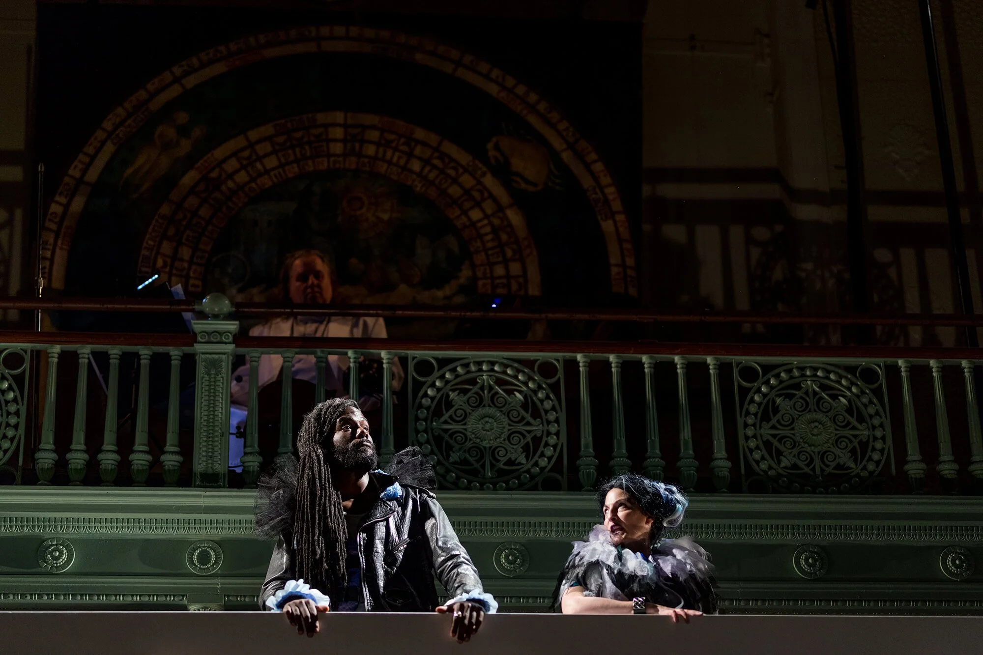Two actors on stage, one with long dreadlocks and a leather jacket, the other with elaborate black and white costume, performing in front of an ornate green railing and a mural of zodiac symbols in the background.