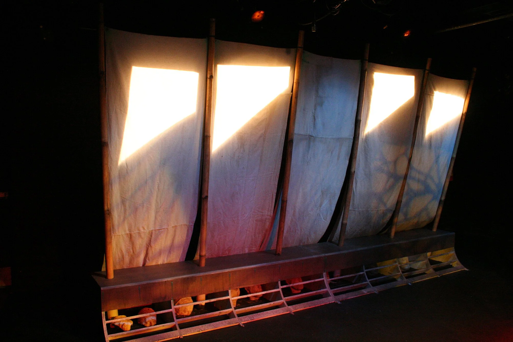 Stage with vertical fabric panels and sunlight streaming through gaps, wooden frame at bottom, set for a performance.