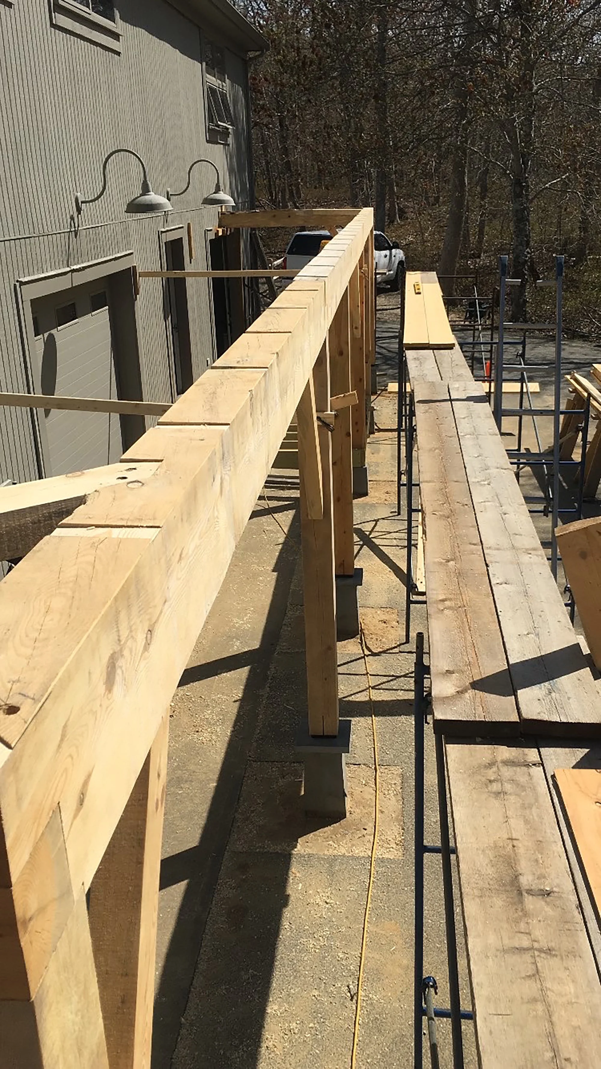 Wooden construction framework with beams and scaffolding outside a building, with a view of trees and parked vehicles in the background.