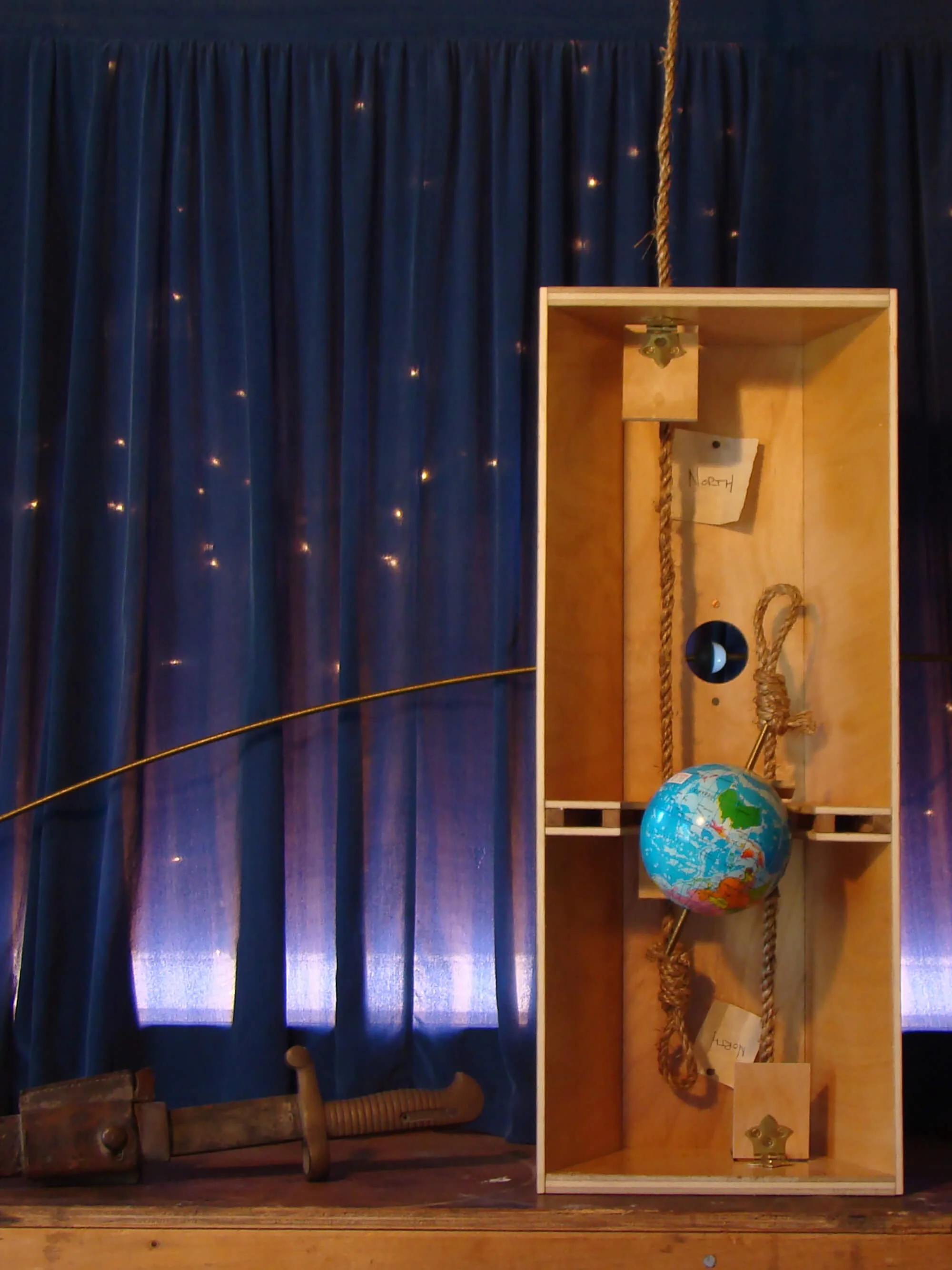 A wooden box with a globe, a rope, and two handwritten notes inside, placed in front of a blue curtain with small lights.