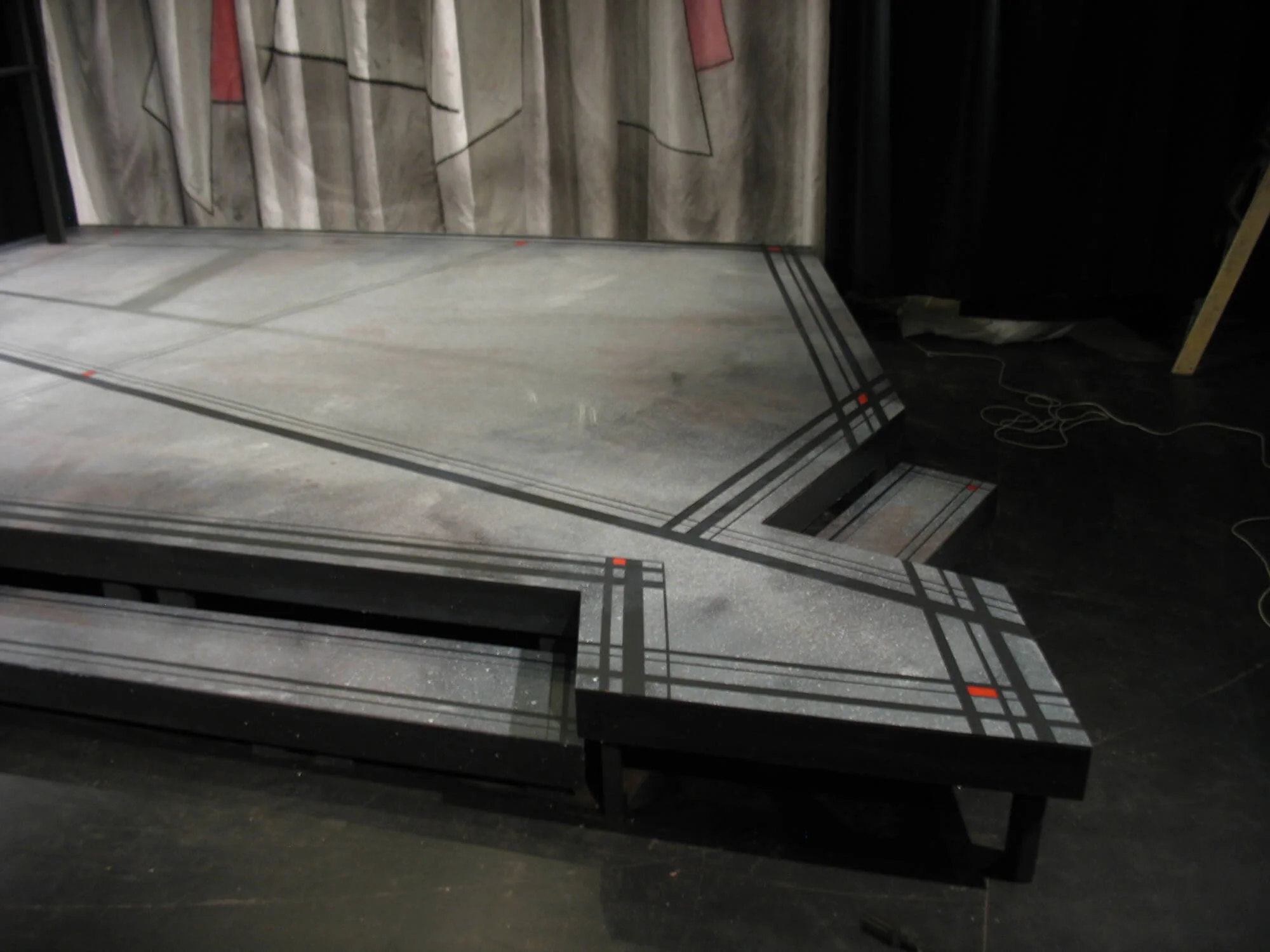 Stage with a patterned gray carpet, partially assembled, in front of a backdrop with abstract designs, surrounded by black curtains and visible electrical cords.