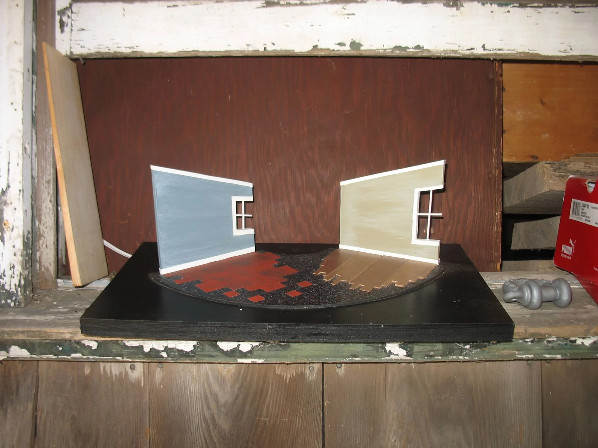 Two small model house walls on a black circular base, one painted blue and the other beige, with a small window on each. The background shows a wooden shelf with various tools and materials.