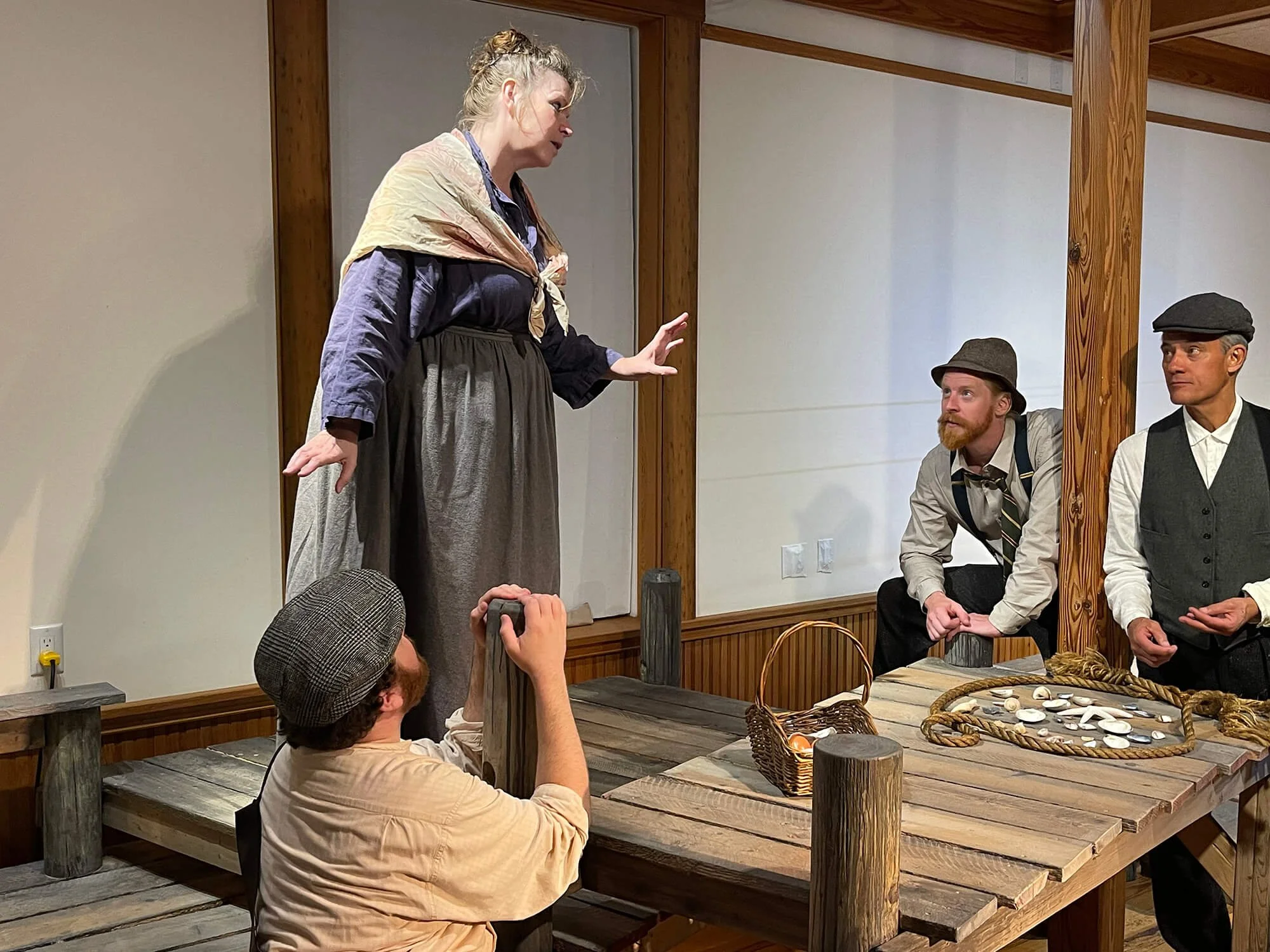 A theatrical performance with four actors on stage. A woman standing and gesturing while three men sit or stand nearby. The set resembles a rustic, old-fashioned setting with a wooden table and props.