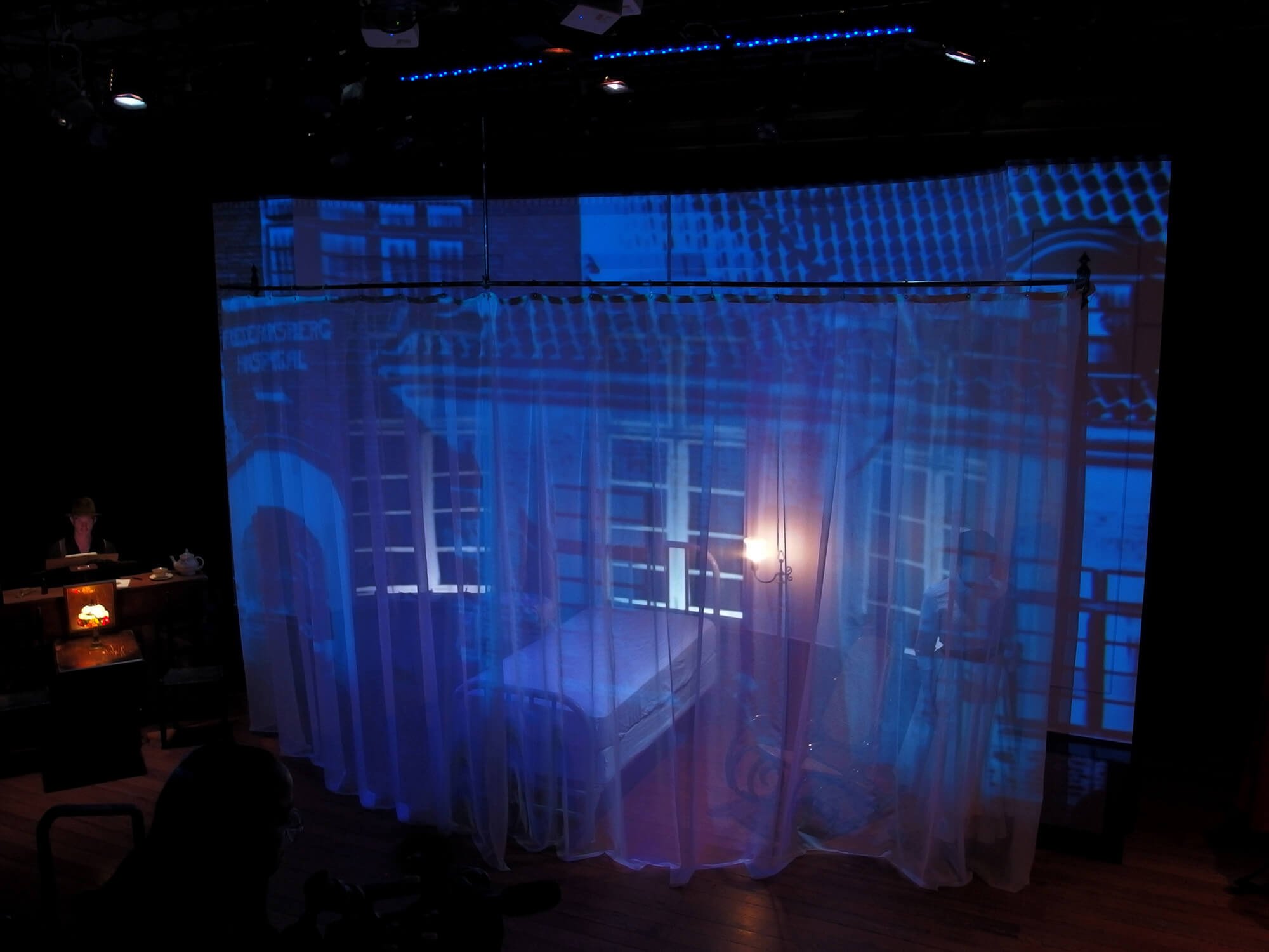 The scene appears to be a theatrical or artistic setup with a bed and outfit behind sheer curtains, illuminated by a soft, warm light. A woman is seen sitting at a desk to the left, and a faint figure is visible behind the curtains. Large window proj