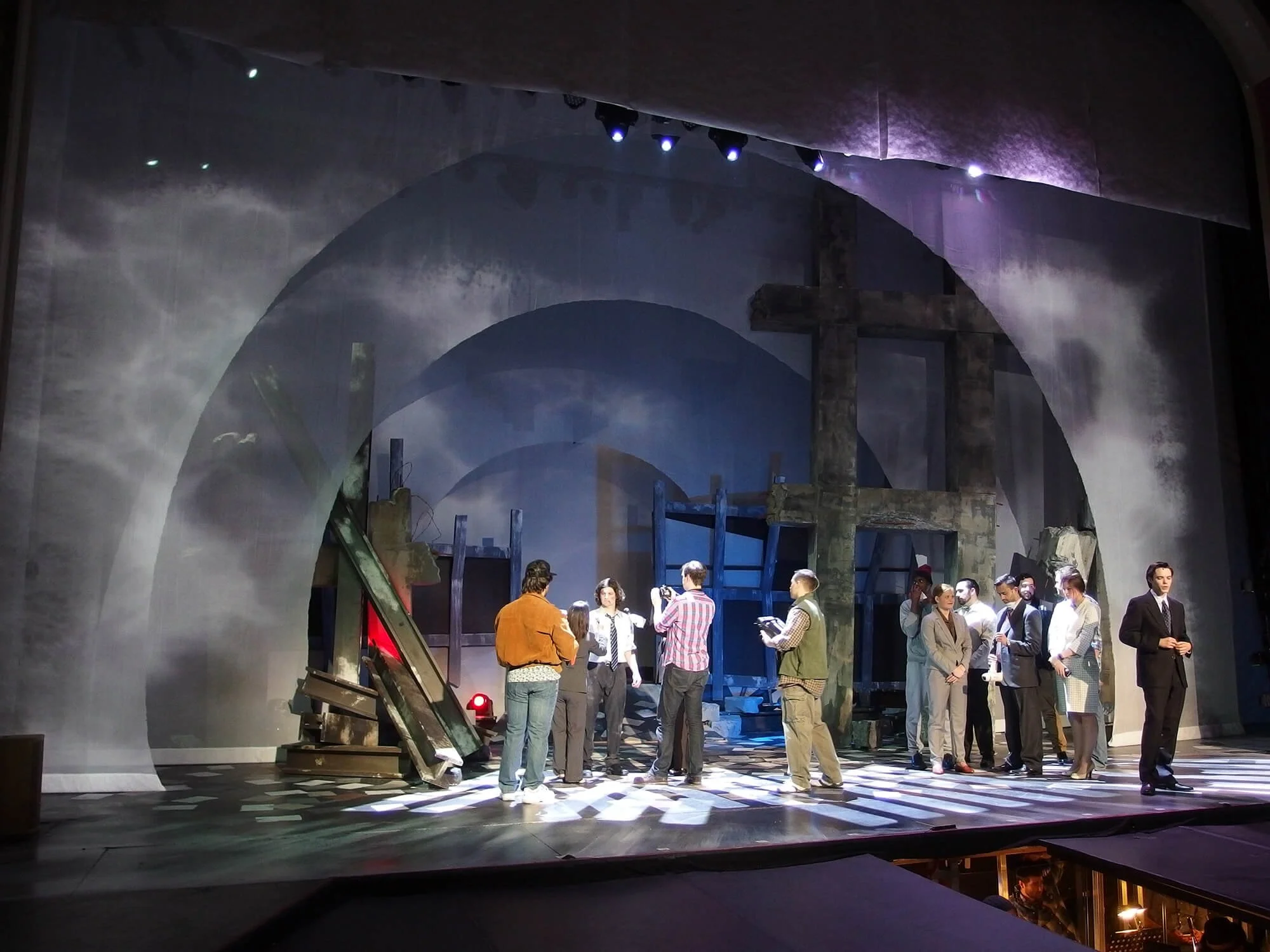 Theatrical stage with actors preparing for a performance, set with large crosses, scaffolding, and dramatic lighting.