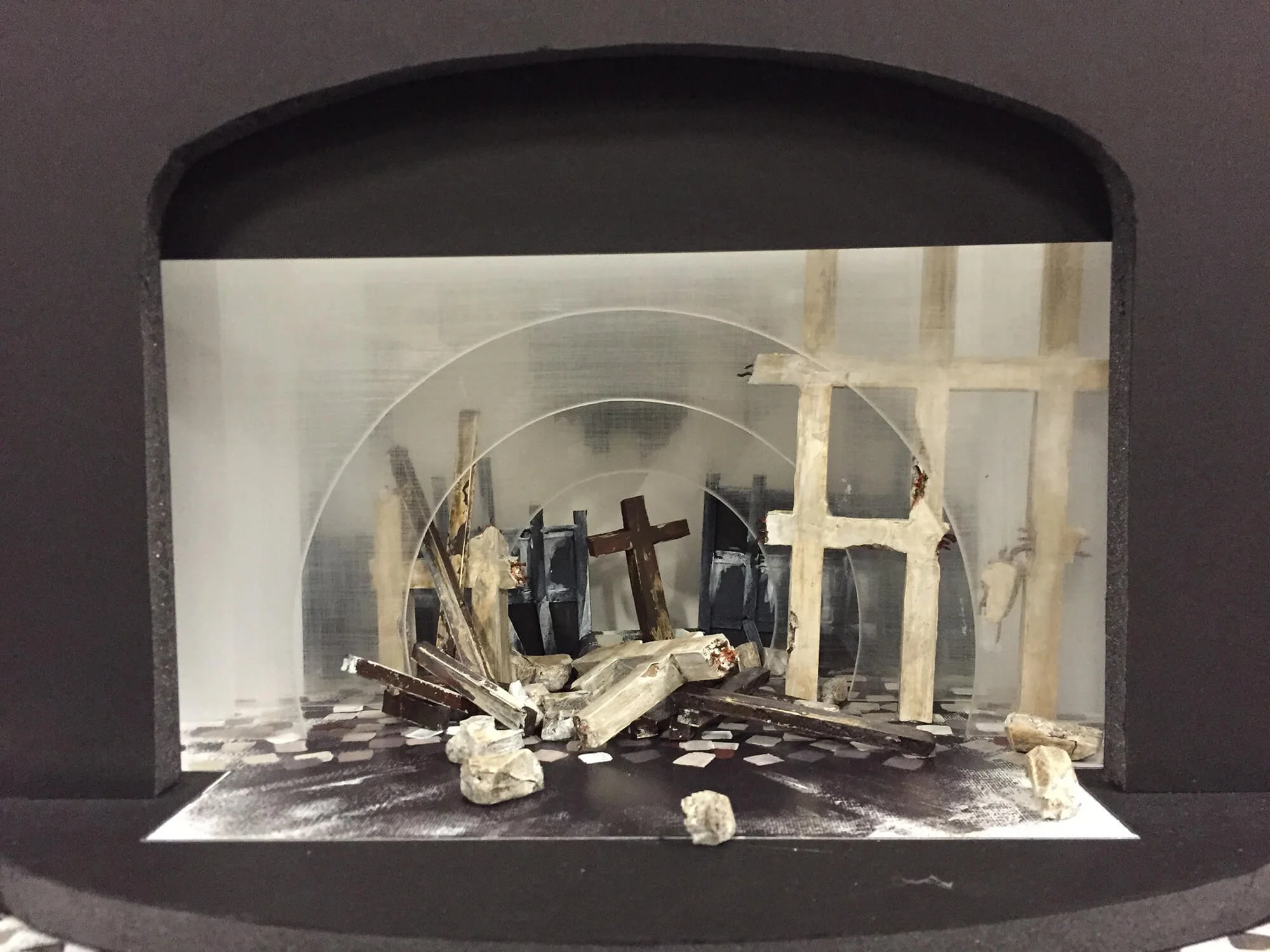 Miniature model of a nativity scene inside a black display case, featuring wooden crosses and scattered rocks, with a background resembling stone or marble.