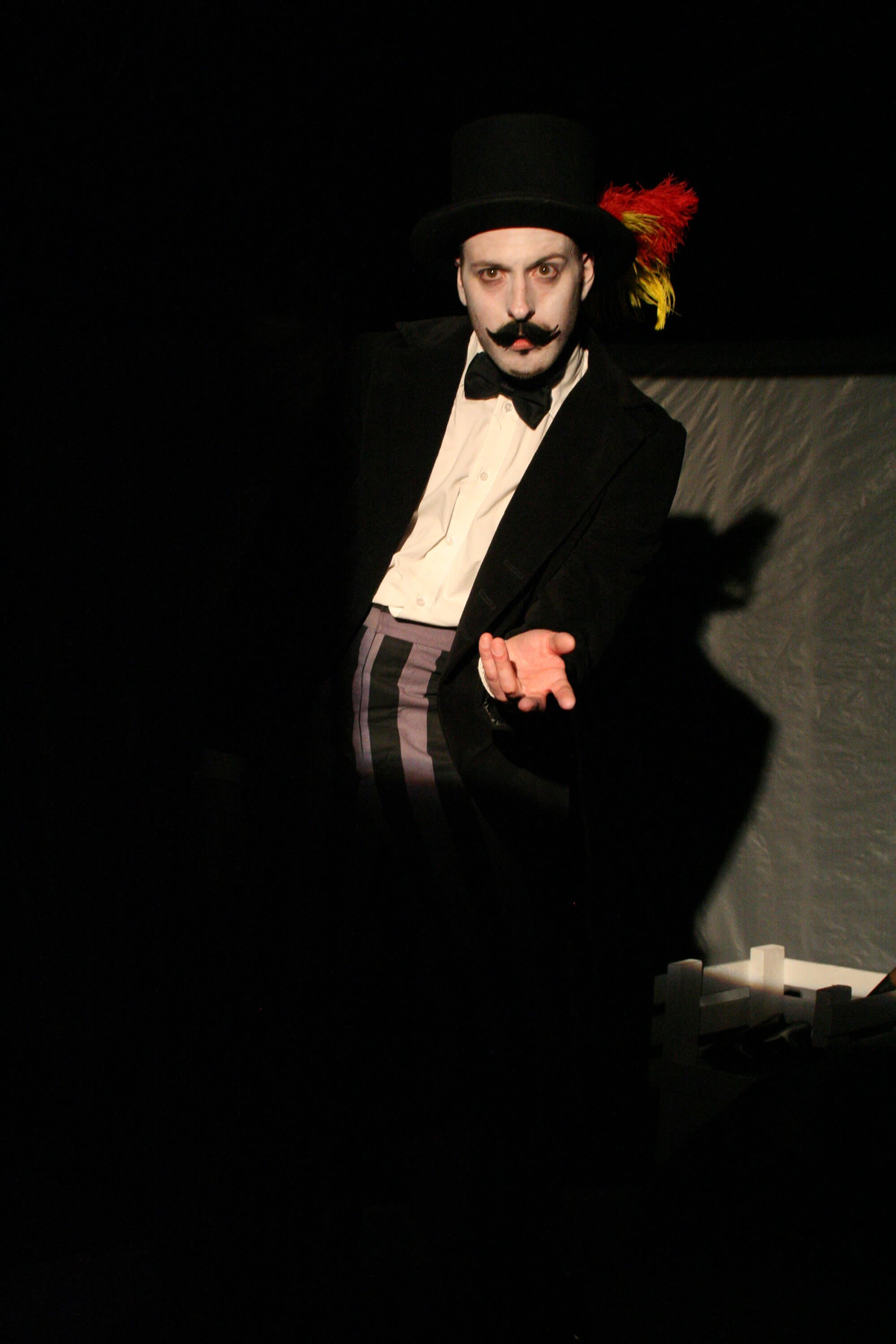A man dressed as a mime clown wearing a black top hat with red and yellow feathers, black suit, white shirt, and black bow tie performing on stage, with dramatic lighting highlighting his face and hand.