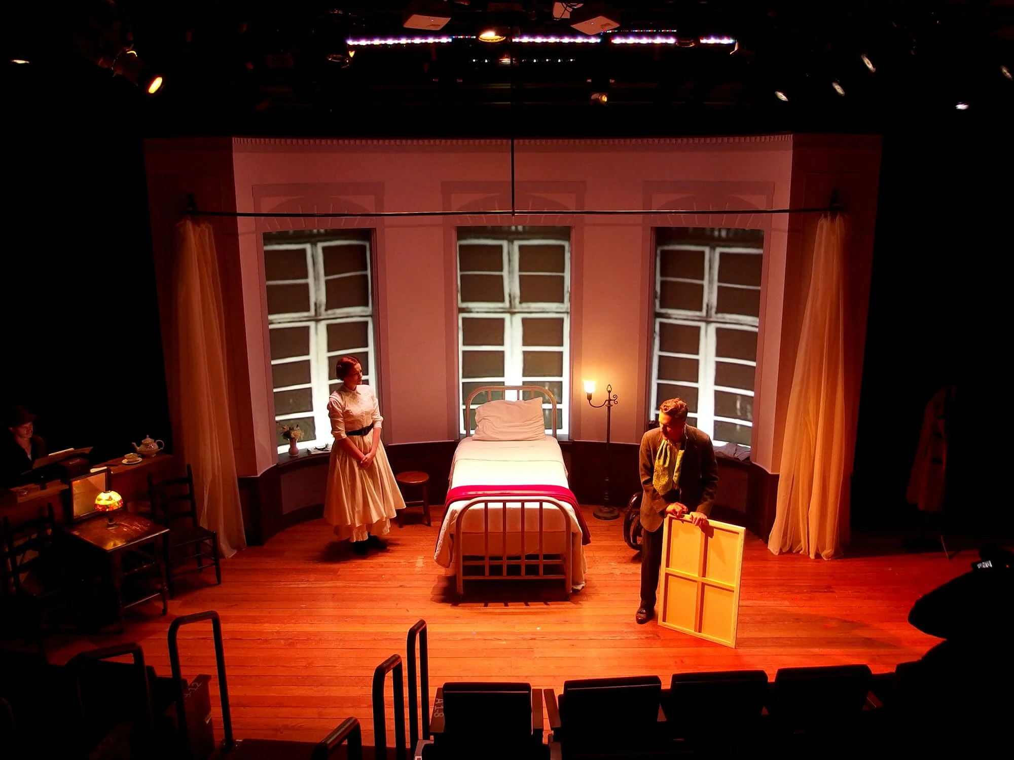 Stage scene of a theater play with three actors in period costumes, a bed in the center, vintage furniture, and a backdrop of windows with white frames.