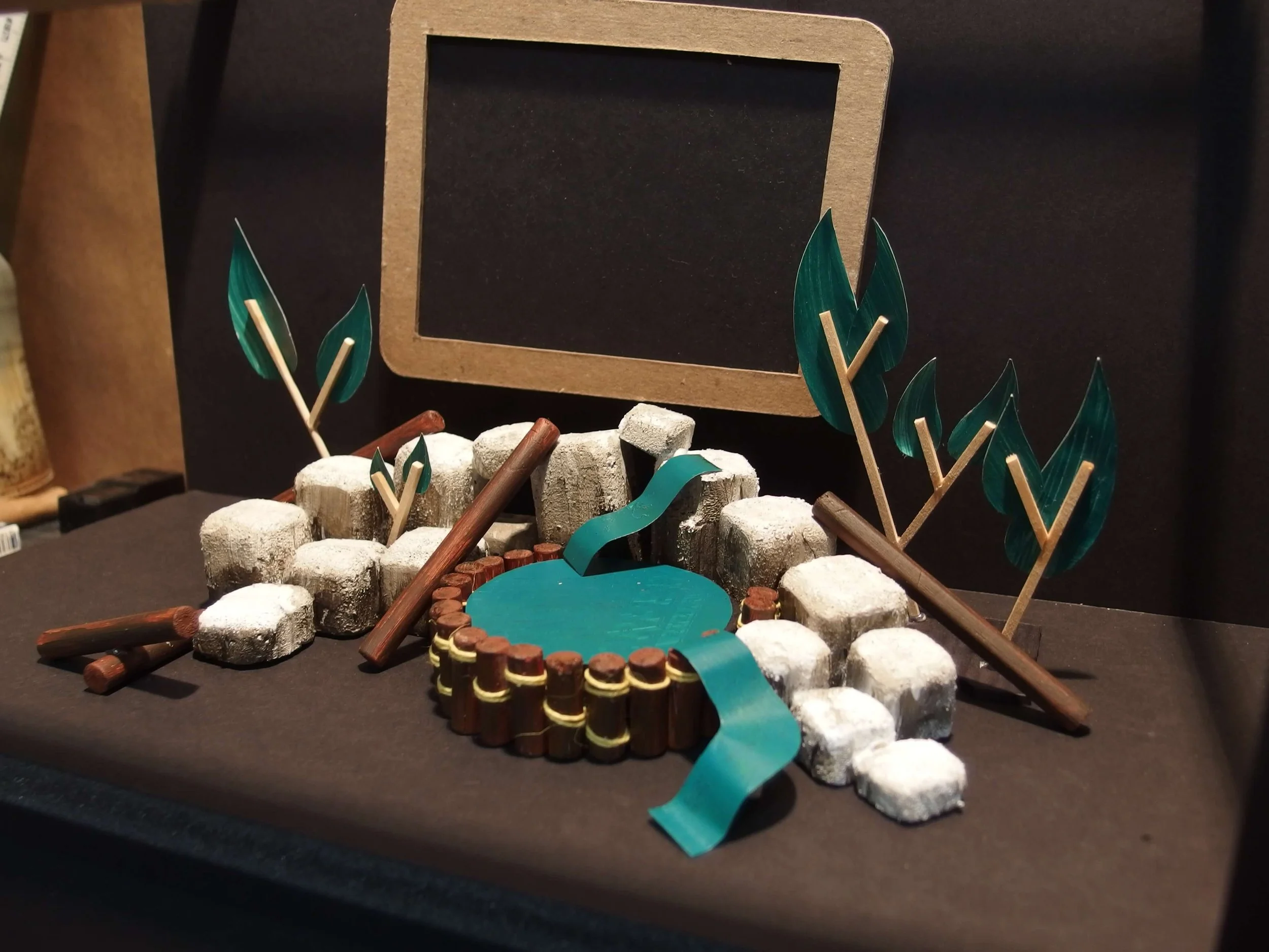 Miniature scene with sugar cubes, wooden sticks, marshmallows, a teal ribbon, and decorative leaves, with a blackboard in the background.