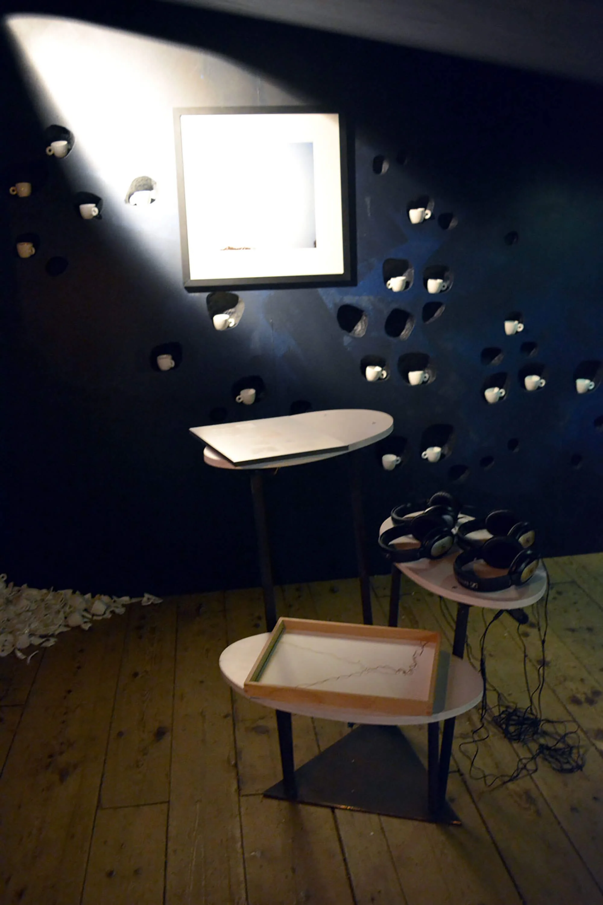 A dark room with a wall decorated with multiple small, white cups placed in holes. A brightly lit, empty square frame is on the wall. There are three small tables, one with a headset, one with a tray, and one holding several headsets. The floor is wo
