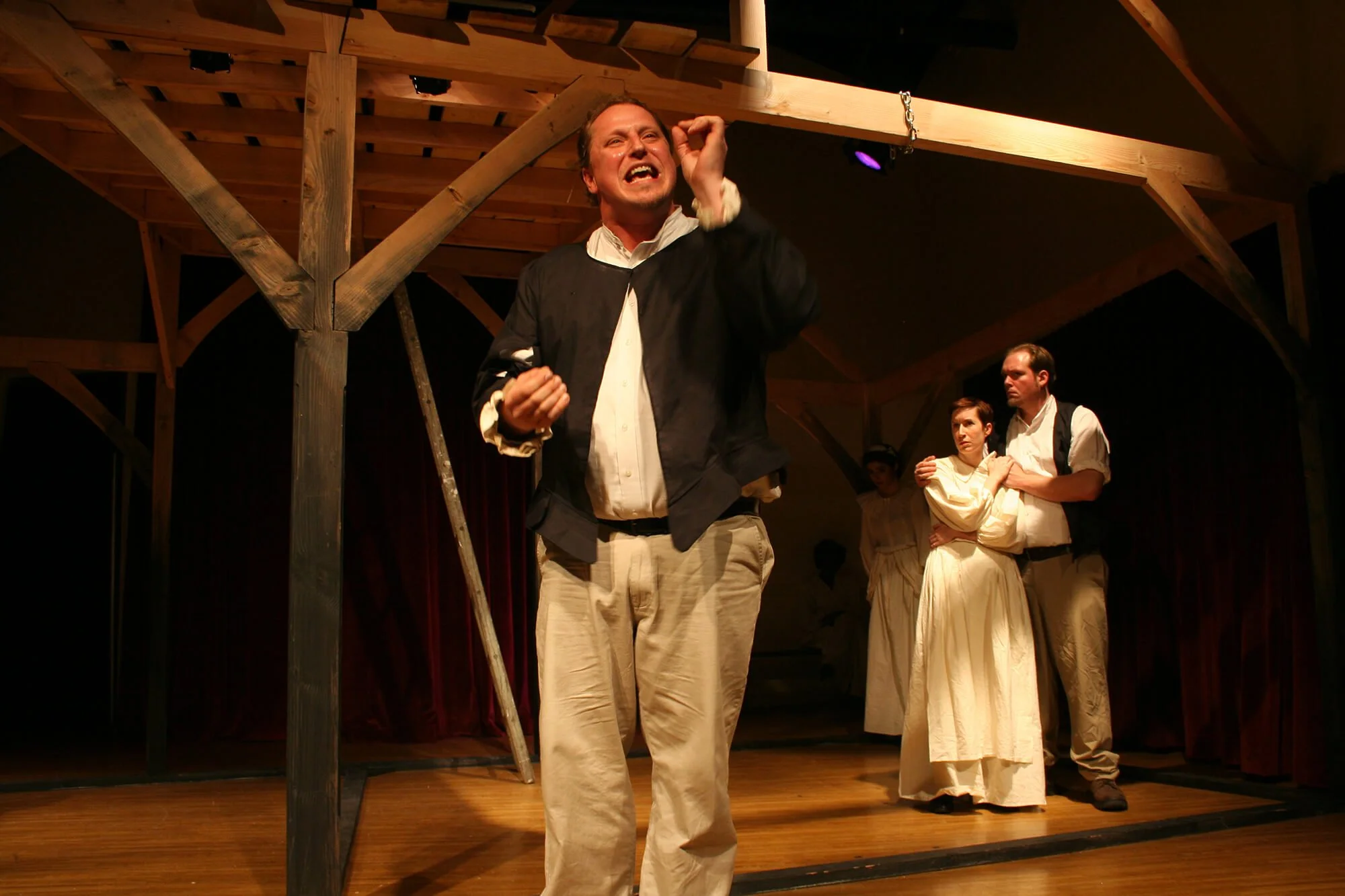 Actors performing in a theatrical play, with a man in the foreground expressing emotion and two actors in the background holding each other.