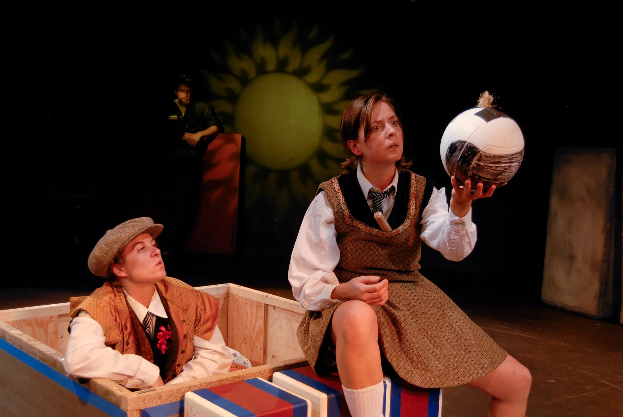 Two young women in vintage costumes perform on stage, one sitting in a wooden box and the other holding a spherical object, with a dark backdrop featuring a yellow sun with radiating rays.