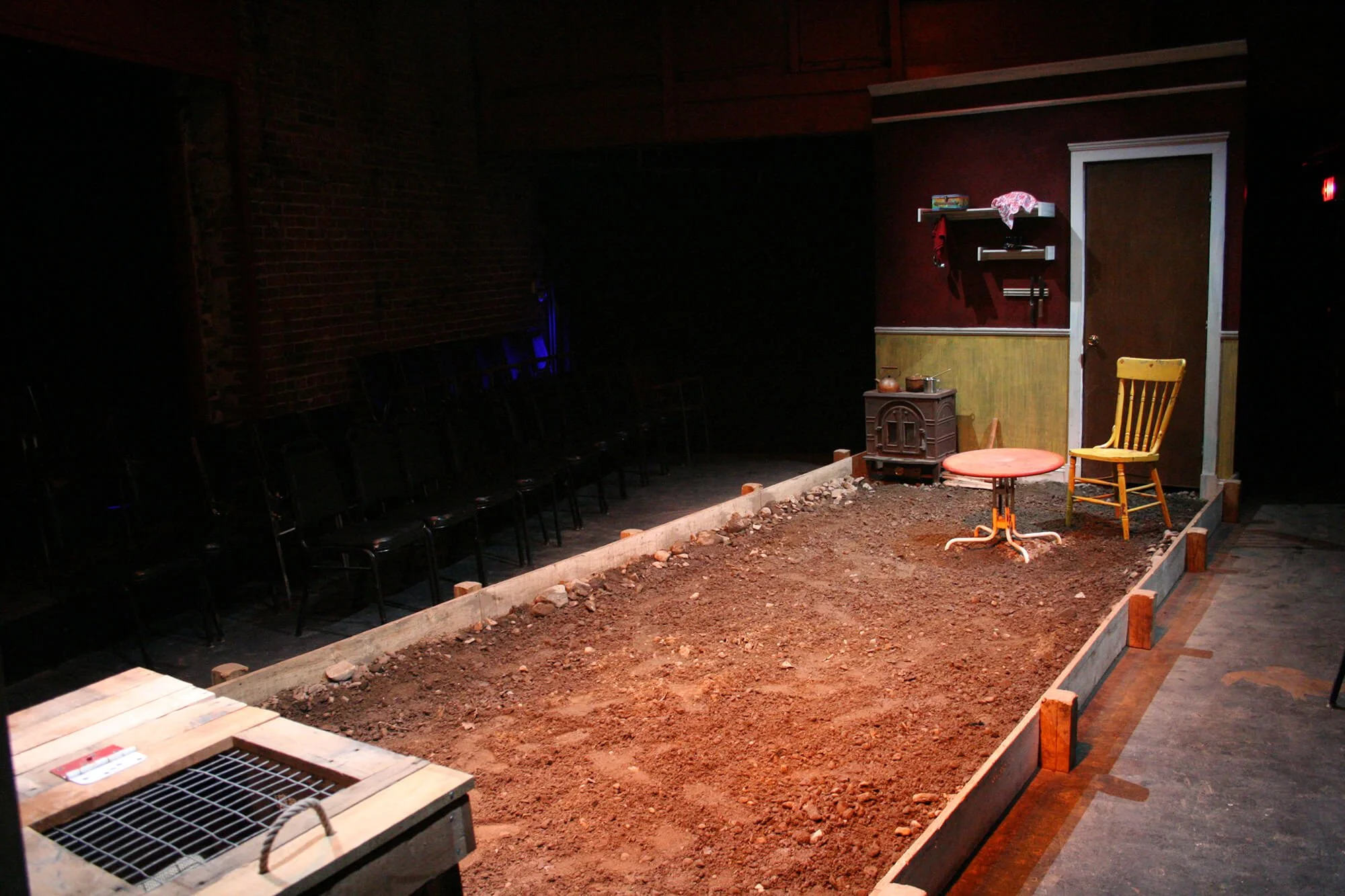 The image shows a small, indoor backyard scene set up as a stage with dirt ground, a small stove, a yellow chair, a small round table, and shelves on the wall. There are black chairs lined up against the wall on the left side of the space.