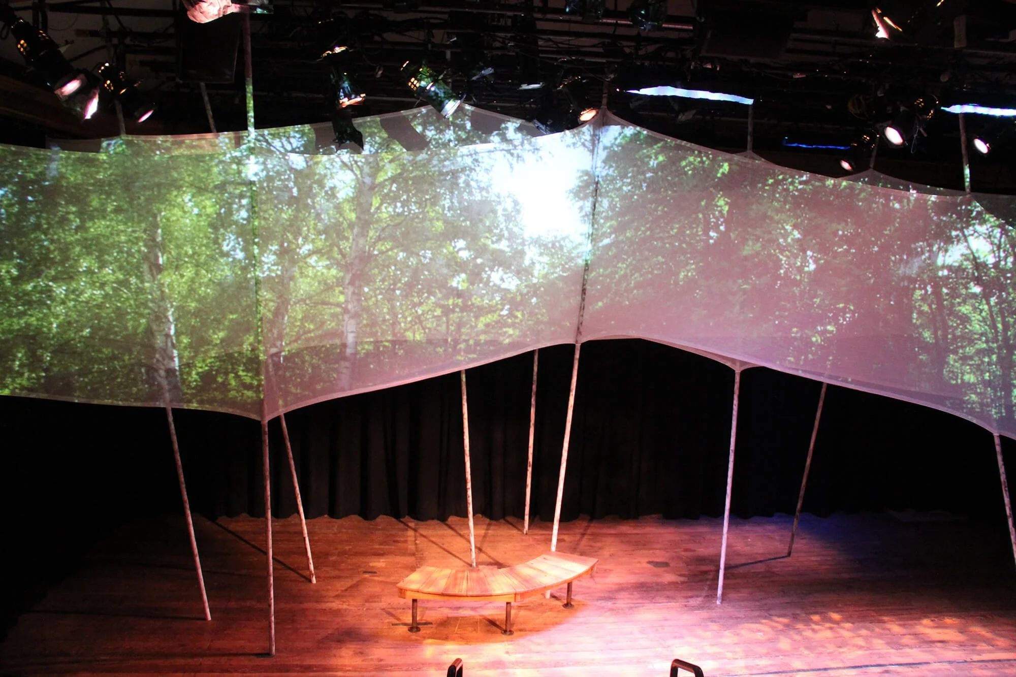 An indoor stage with a curved backdrop displaying an outdoor forest scene with green trees and sunlight. The stage has a small semi-circular wooden bench and is lit by several stage lights mounted above.