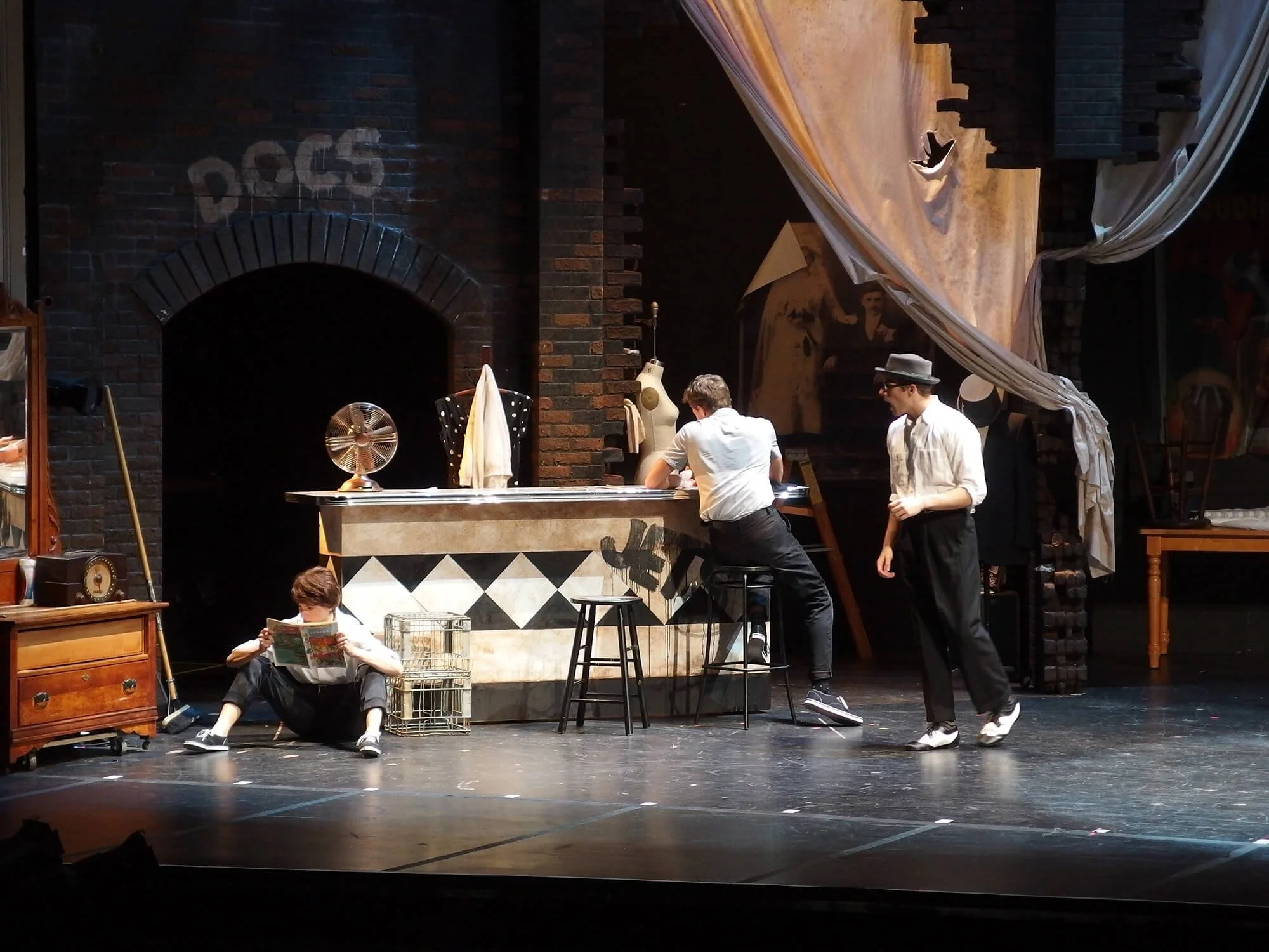 Theatrical stage scene with four actors, set designed as an apartment or flat, with brick walls, a counter, and a large curtain. One young man sits on the floor reading a newspaper, another leans on the counter, and two others stand nearby, engaging 